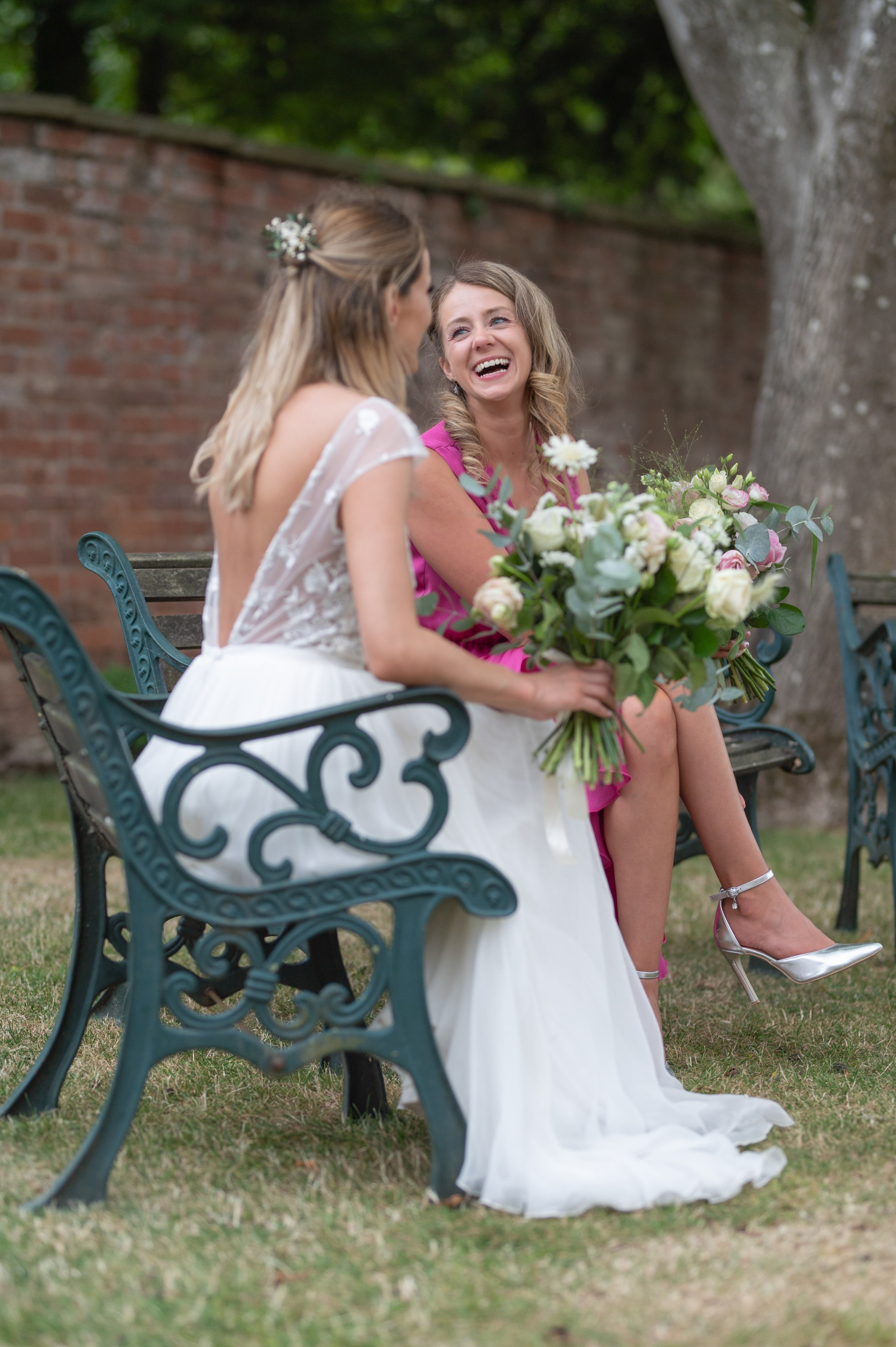 Bride laughing with a bridesmaid in pink, seated together on a garden bench with their bouquets