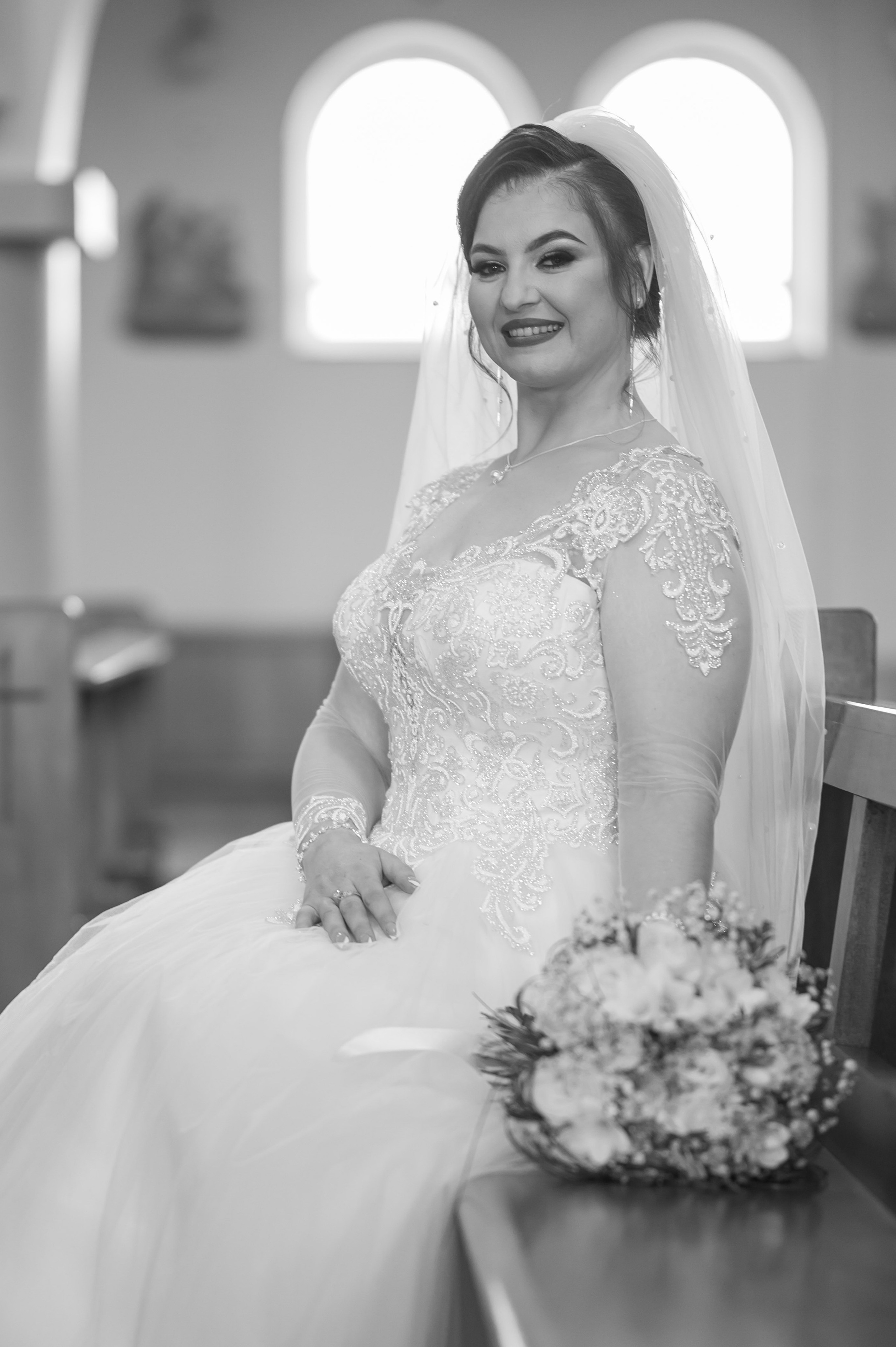 Bride seated in the church, bouquet in hand, beneath arched windows, black and white