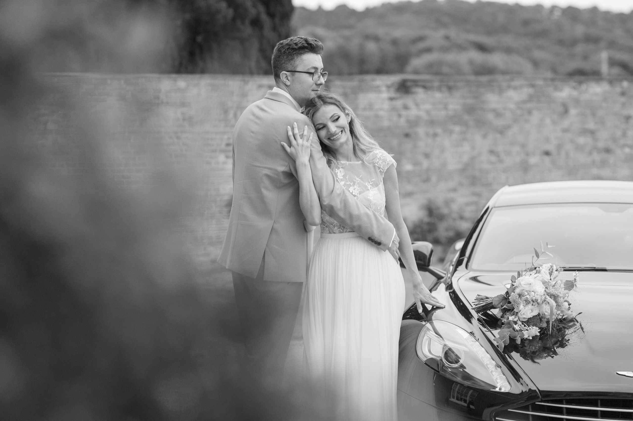 Bride and groom by the Aston Martin wedding car, bouquet resting on the bonnet (black and white)