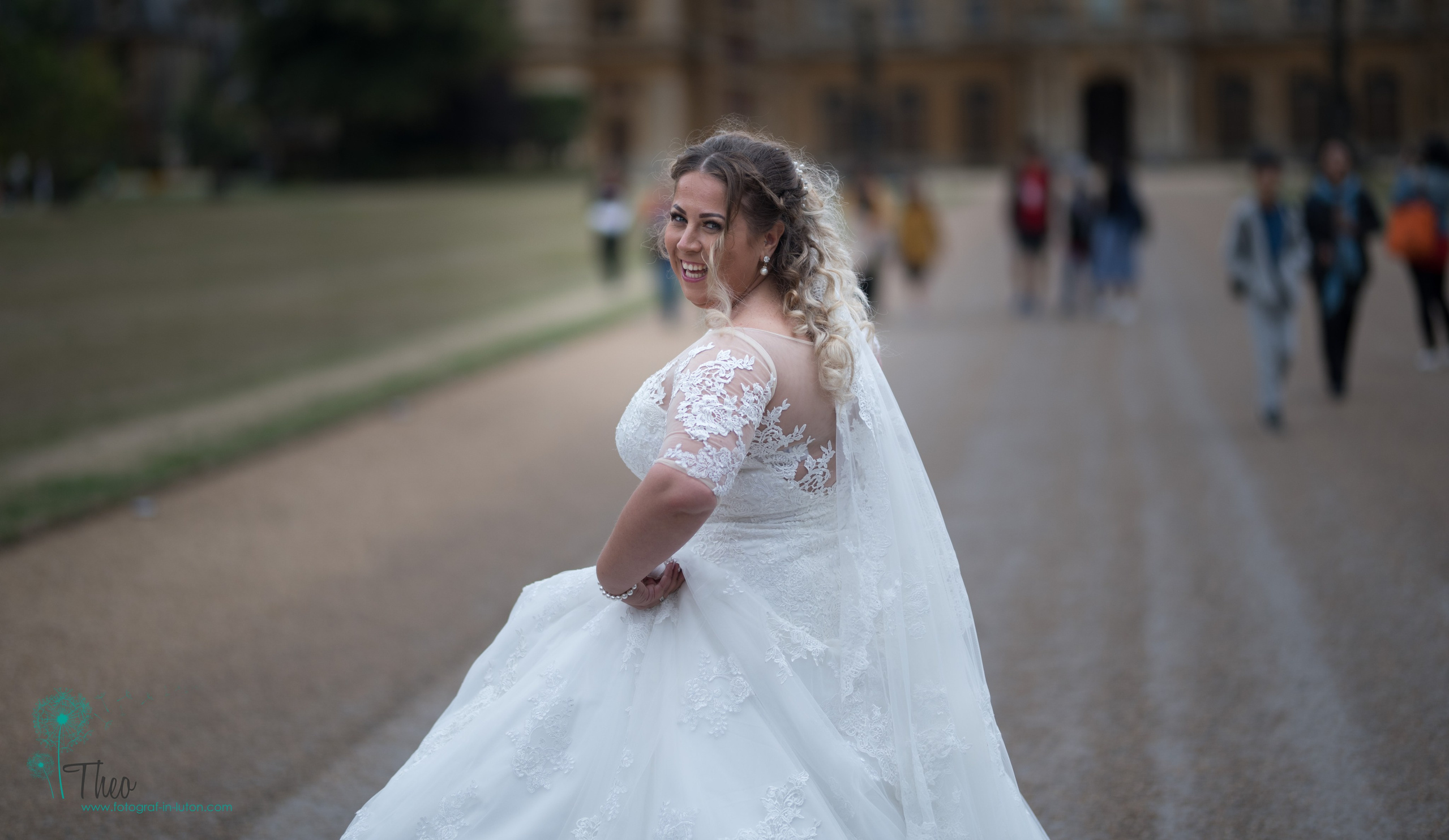 The bride glances back over her shoulder with a joyful smile as she twirls on the drive at Waddesdon Manor, other visitors walking past in the background