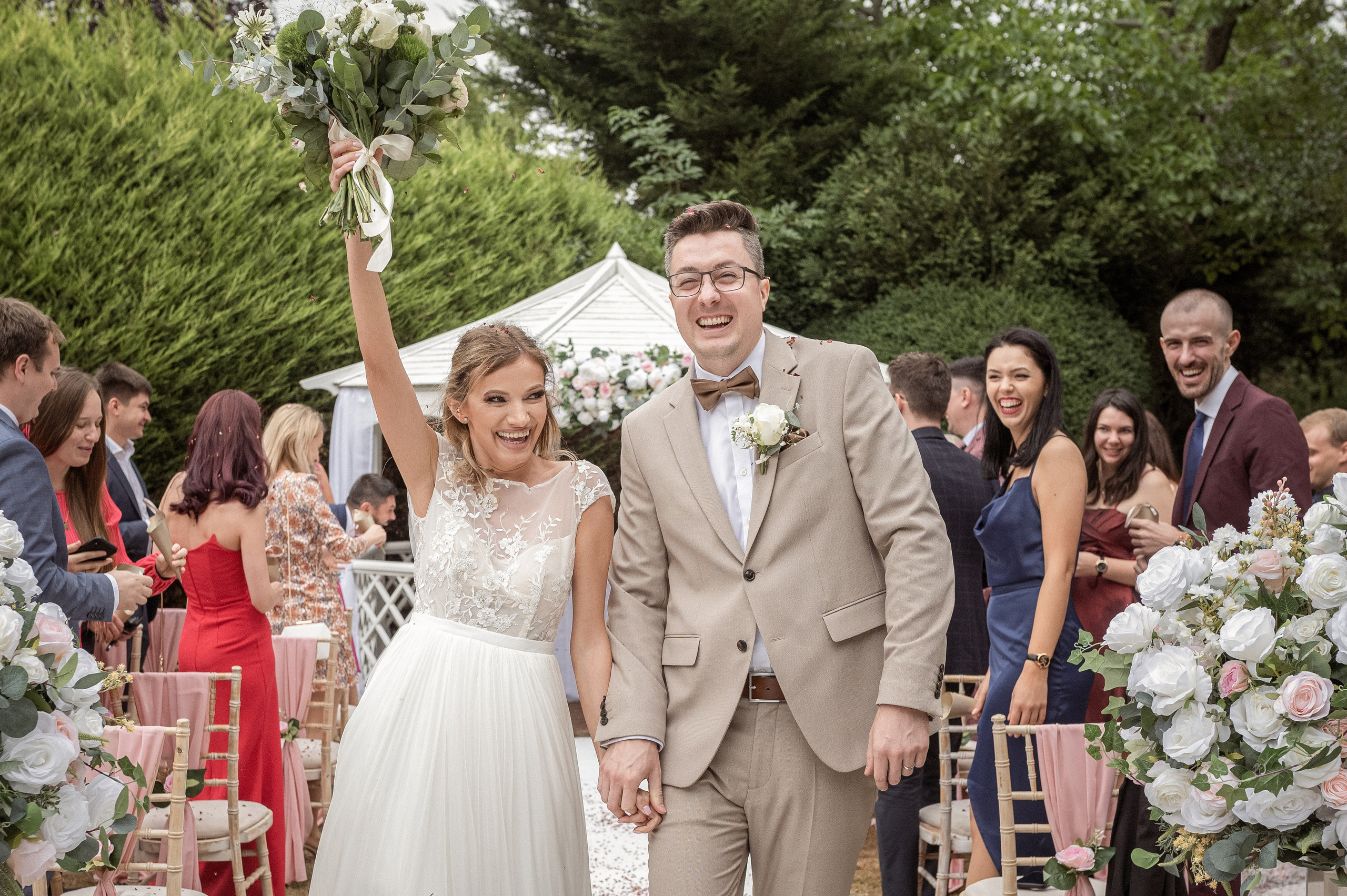 Bride and groom laughing during their confetti walk, bouquet raised, guests celebrating either side
