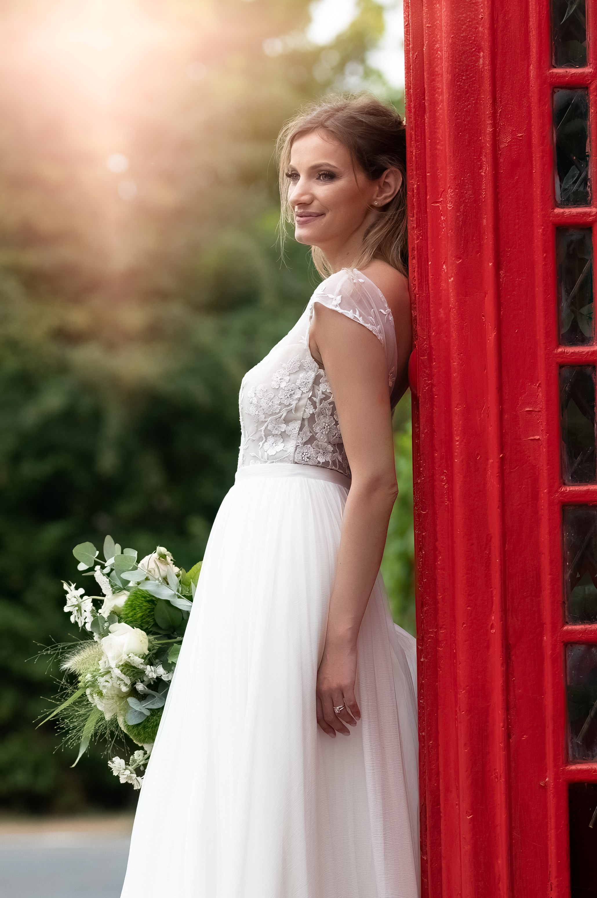 Bride leaning against the red phone box at golden hour, smiling into the distance with her bouquet