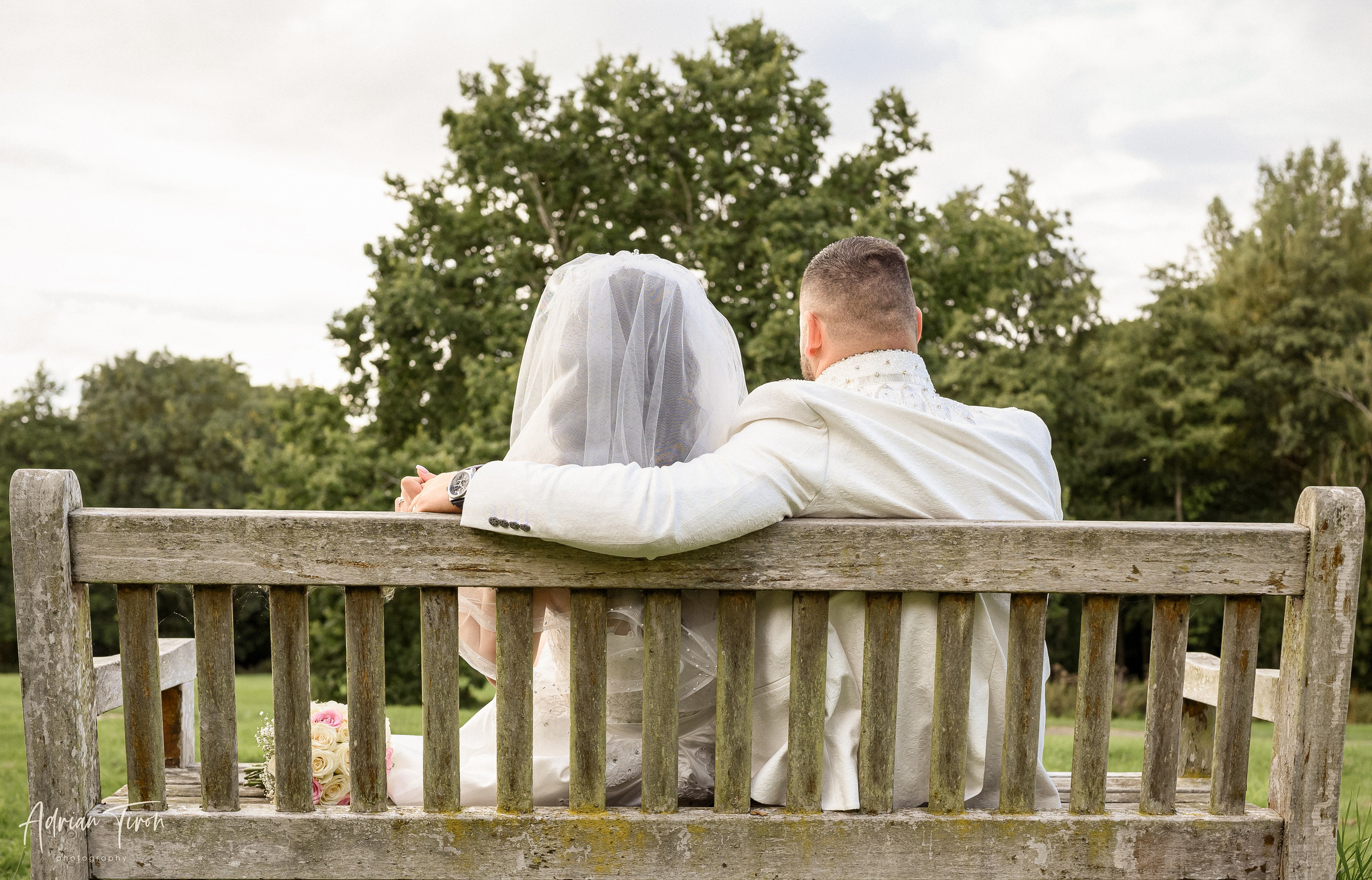 Bride and groom sitting together on a wooden bench, seen from behind, surrounded by trees