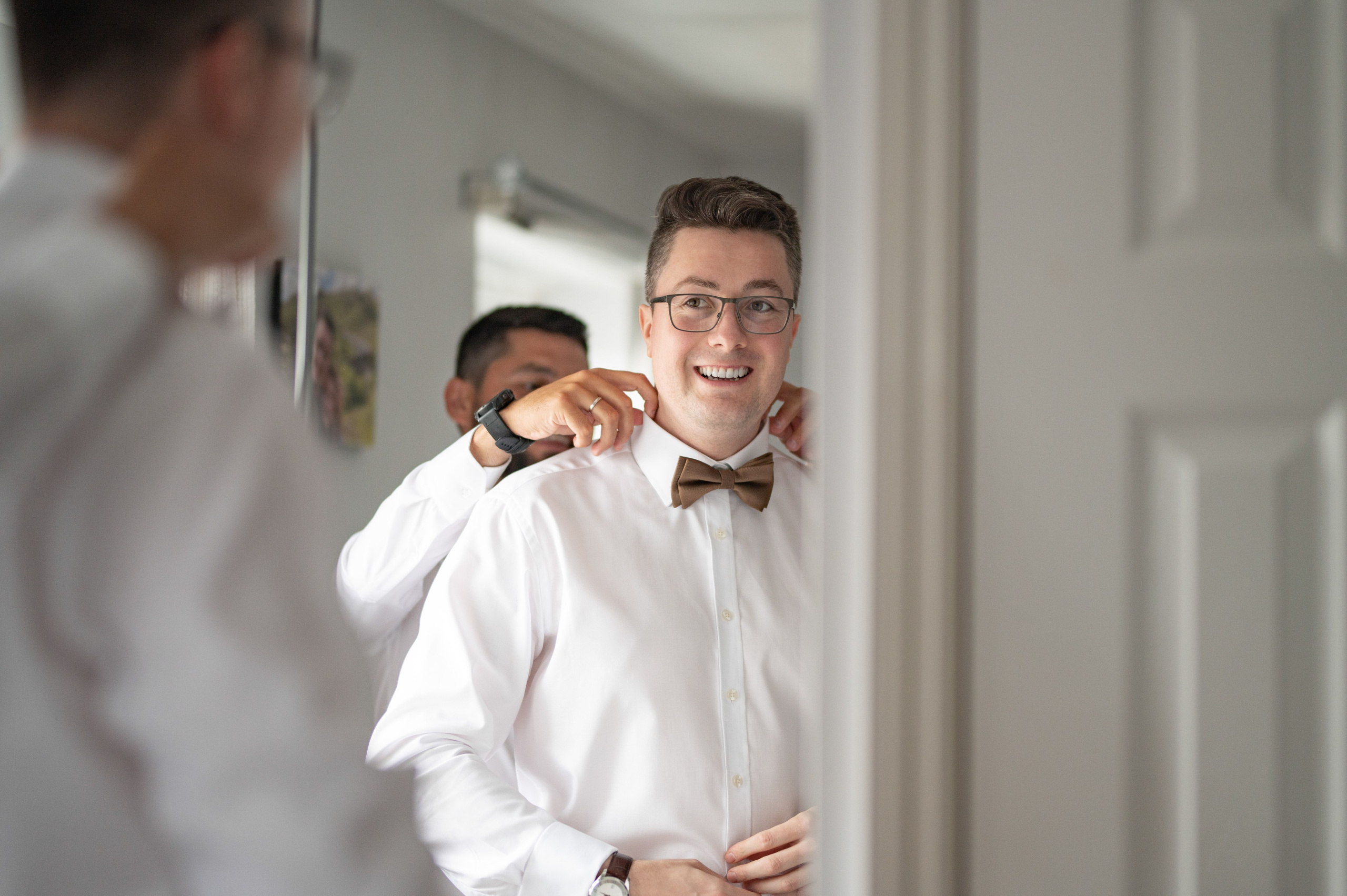 Groom all smiles as his tan bow tie is fastened by a groomsman