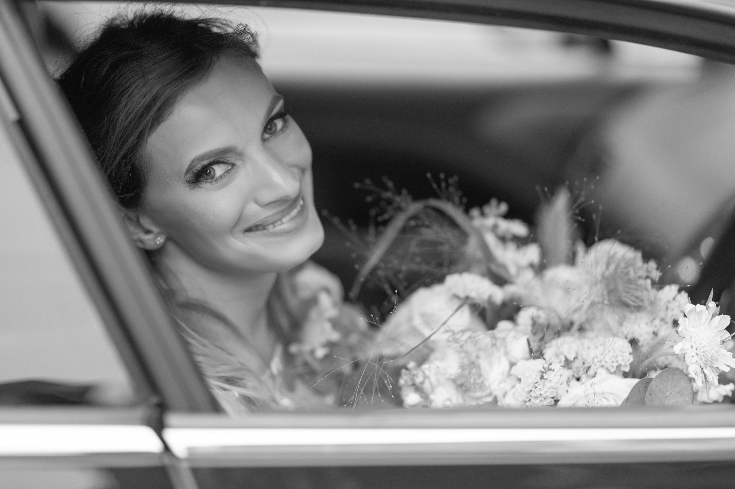 Bride smiling through the car window with her bouquet beside her (black and white)