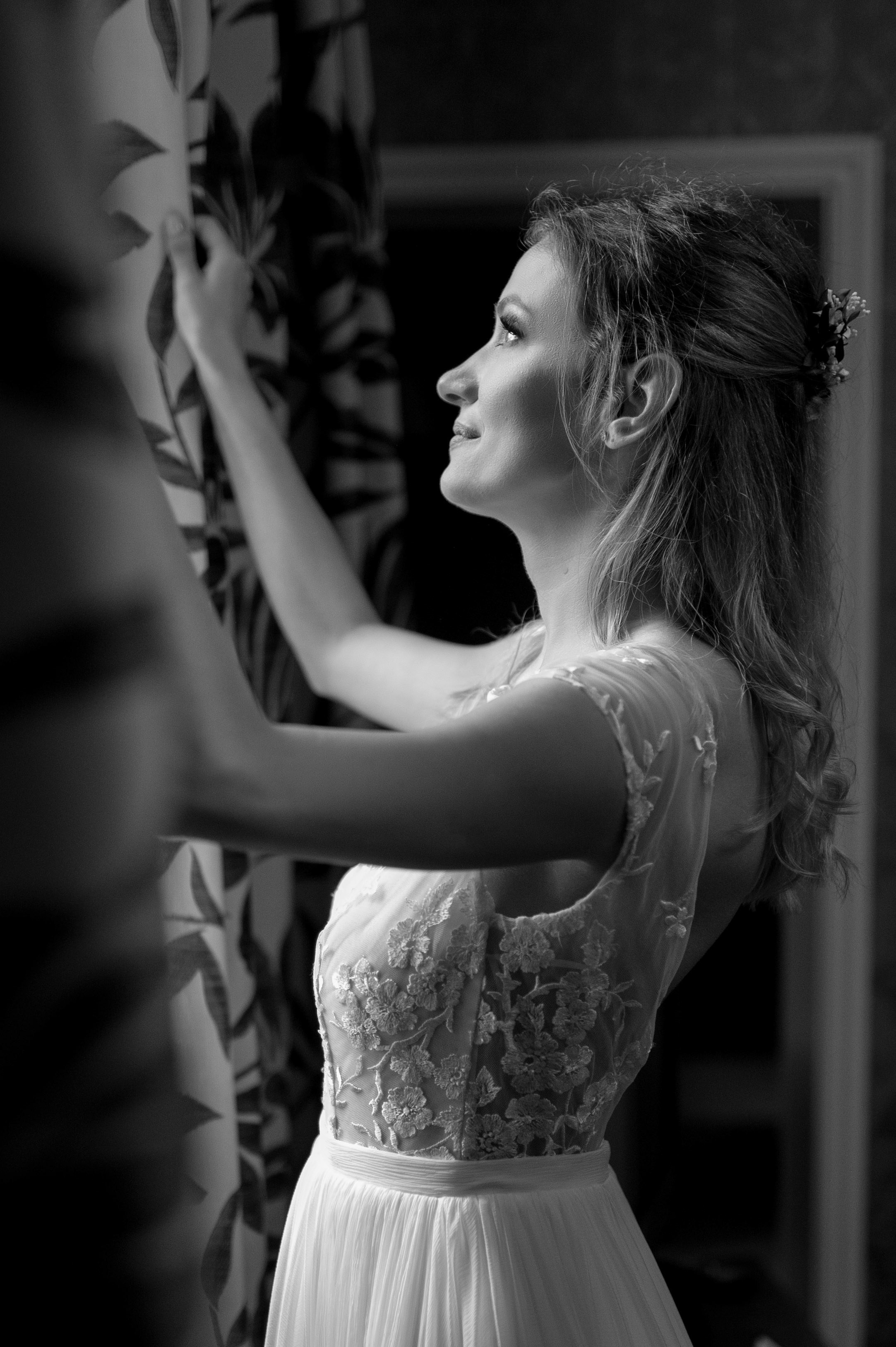 Bride in profile smiling towards the light, holding back the curtain (black and white)