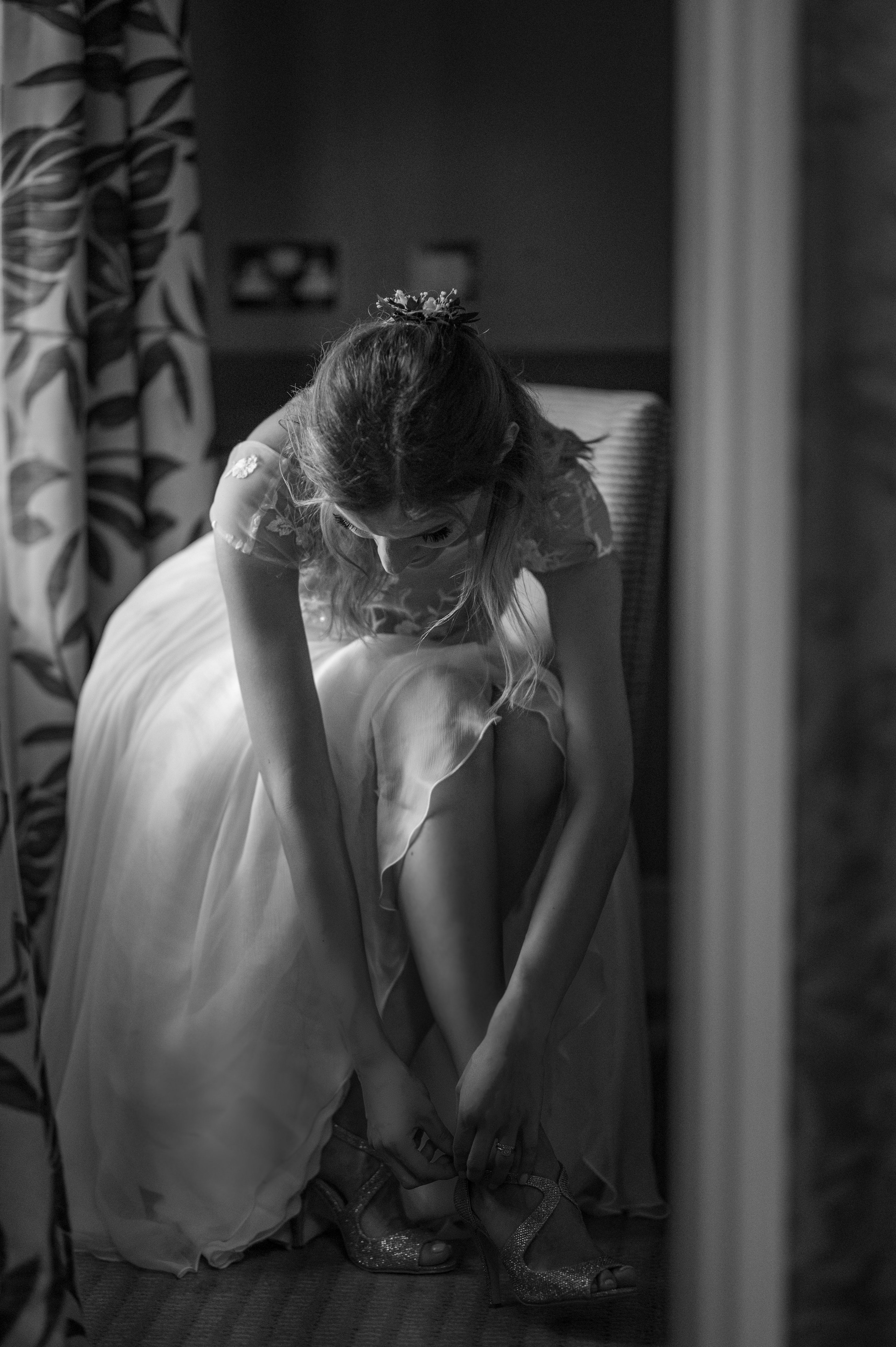 Bride fastening the straps of her sparkly wedding shoes, seated by the window later in the morning (black and white)
