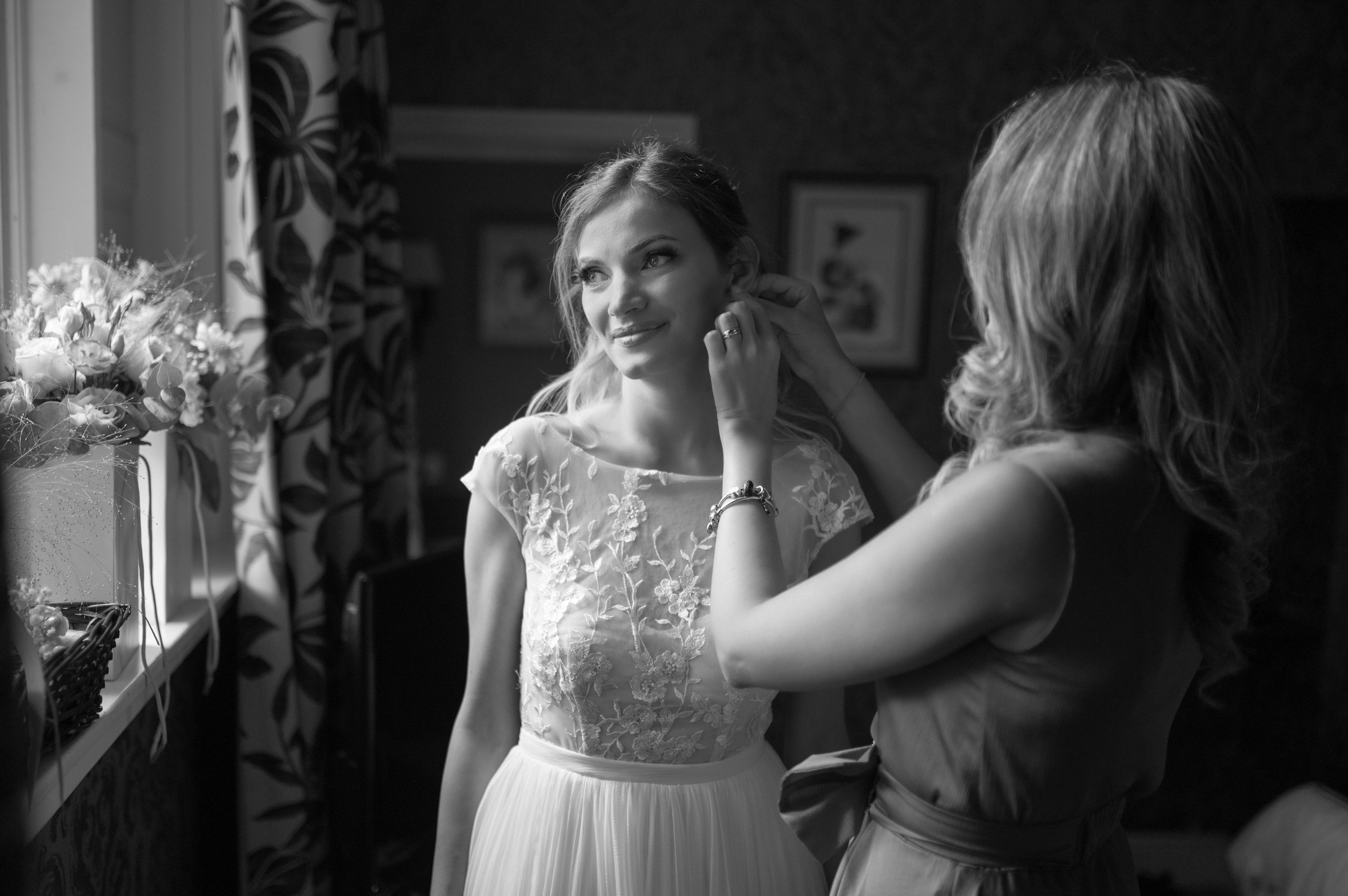 Bridesmaid fastening the bride's earring as she smiles, flowers on the windowsill (black and white)