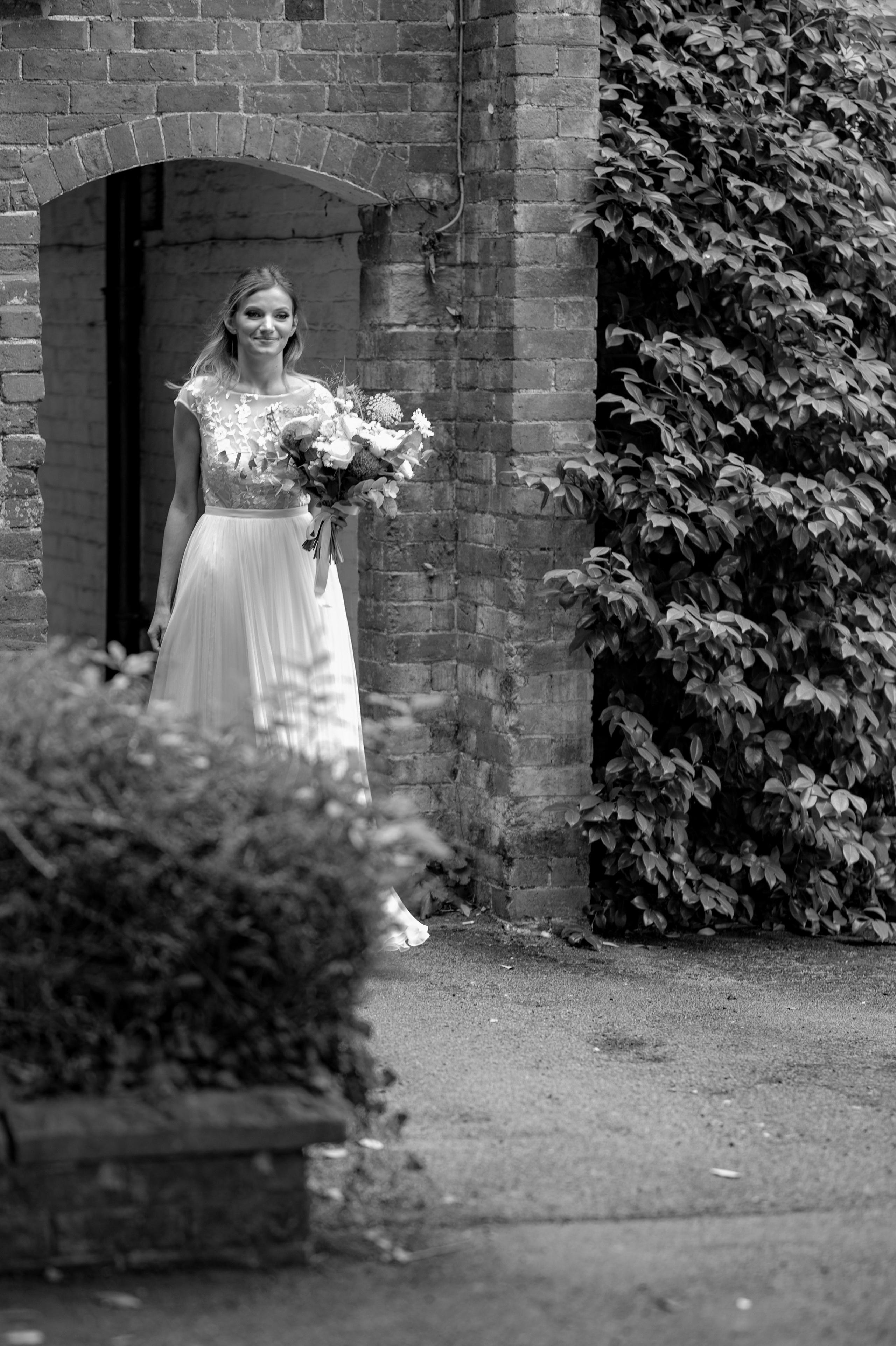 Bride smiling in a brick archway doorway holding her bouquet (black and white)