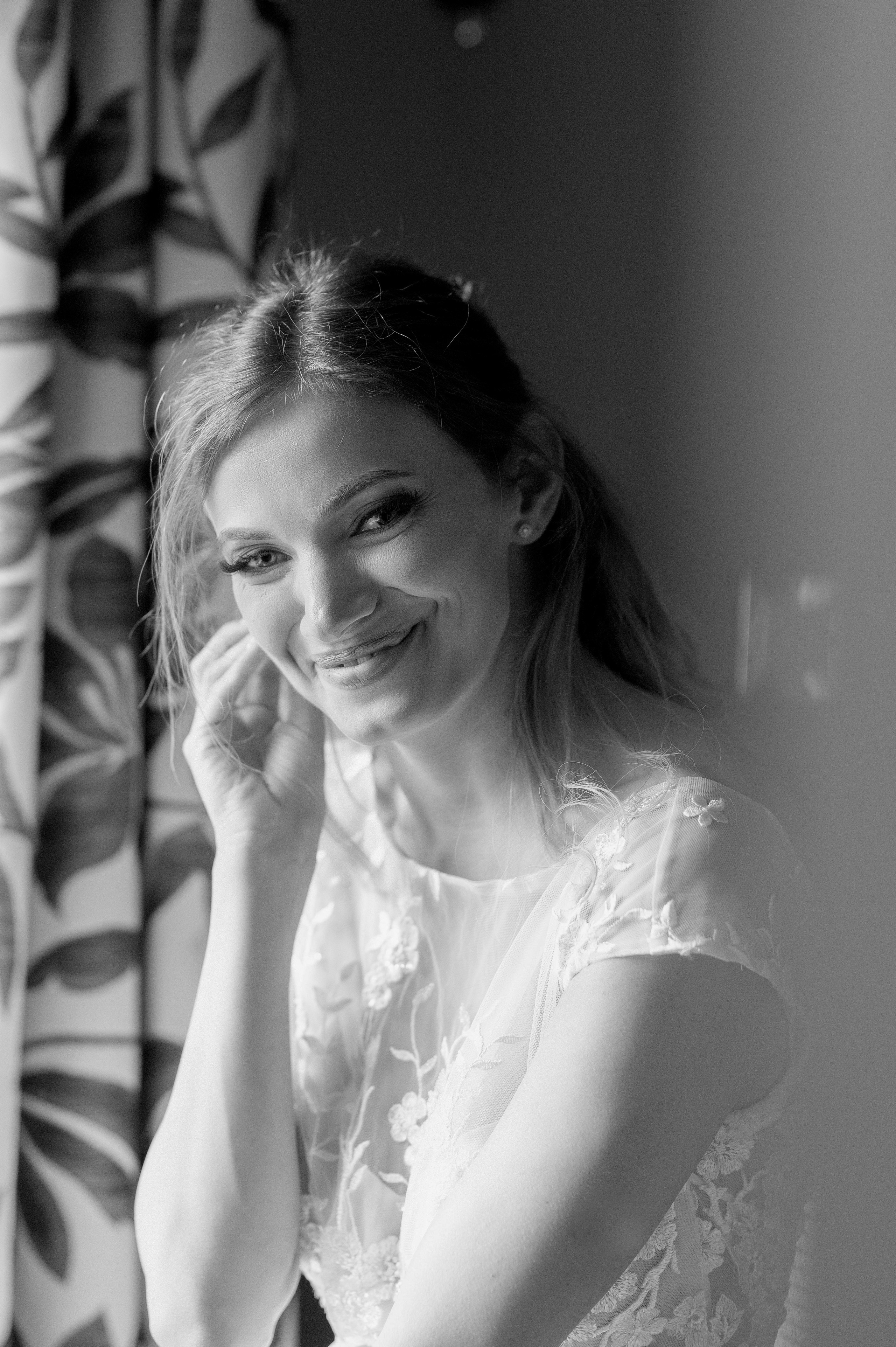 Bride smiling and fastening her earring by the window, soft light (black and white)