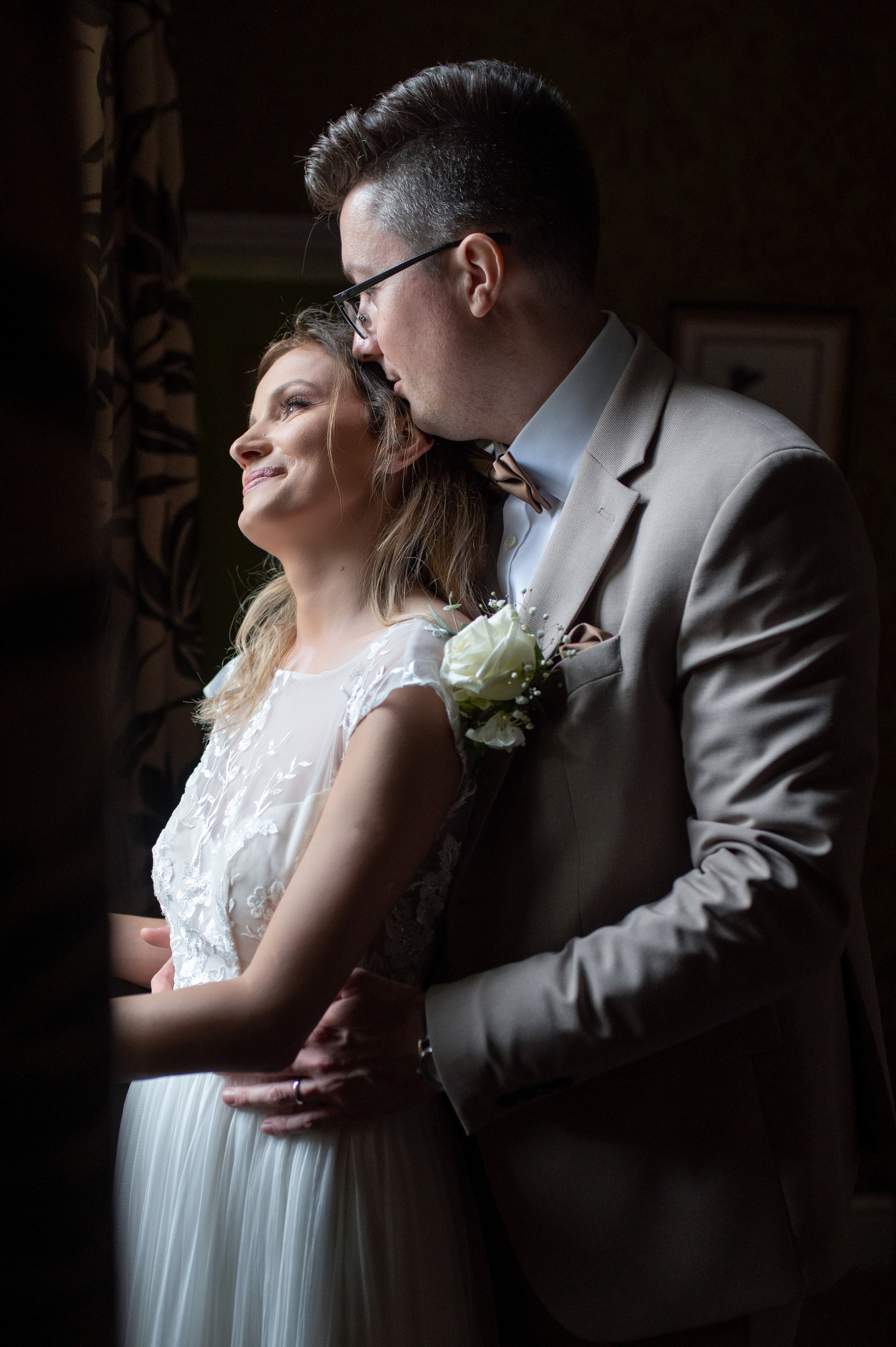 Bride and groom embracing by the window, both looking out with soft natural light
