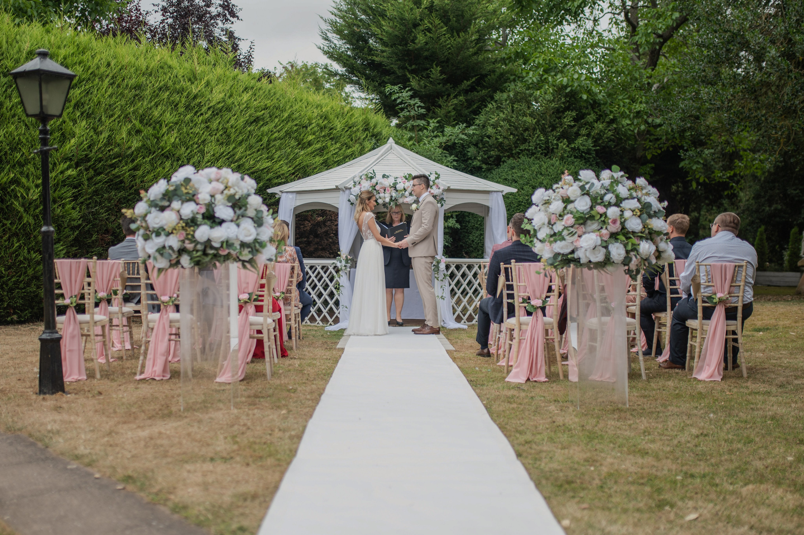 Bride and groom exchanging vows under the white gazebo at Charlecote Pheasant Hotel, pink and white floral aisle decor