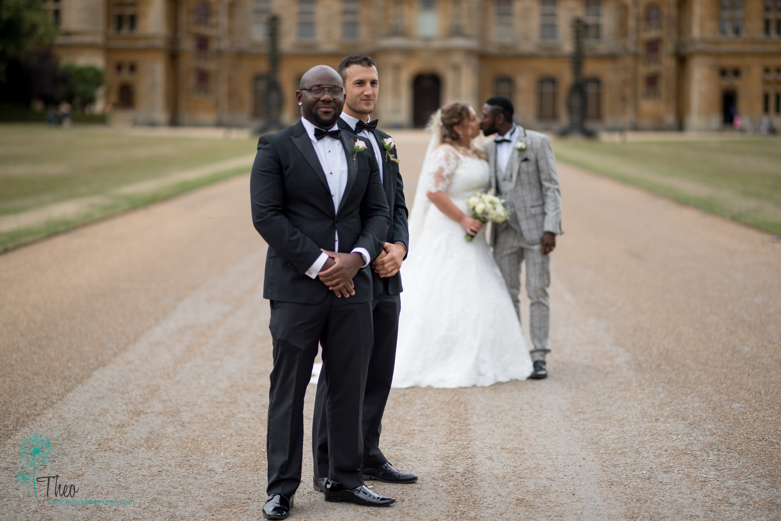 Groomsmen in black tuxedos stand sharply in focus while the bride and groom share a soft kiss behind them at Waddesdon Manor