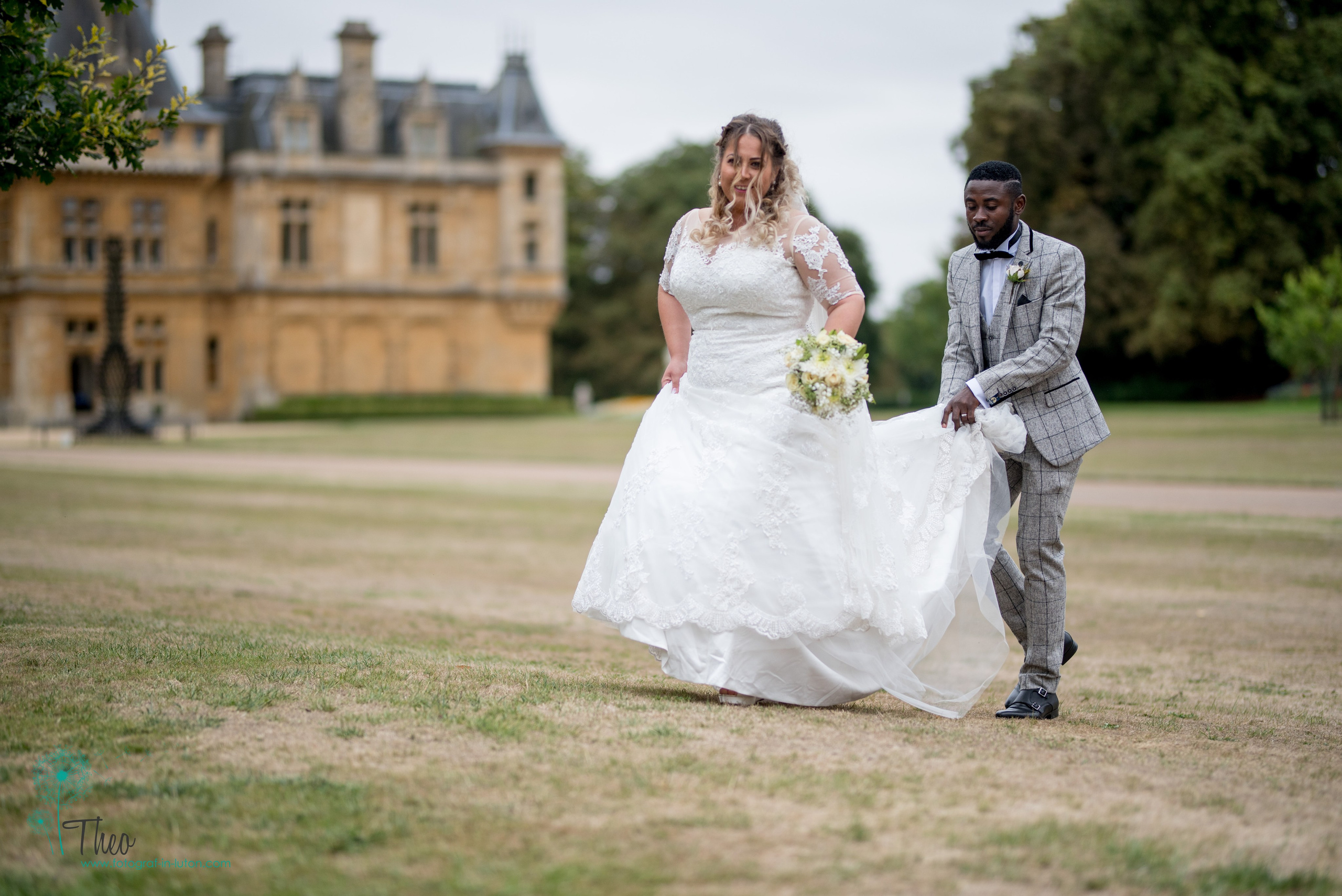 The groom carefully carries his bride's train as they walk across the lawns at Waddesdon Manor