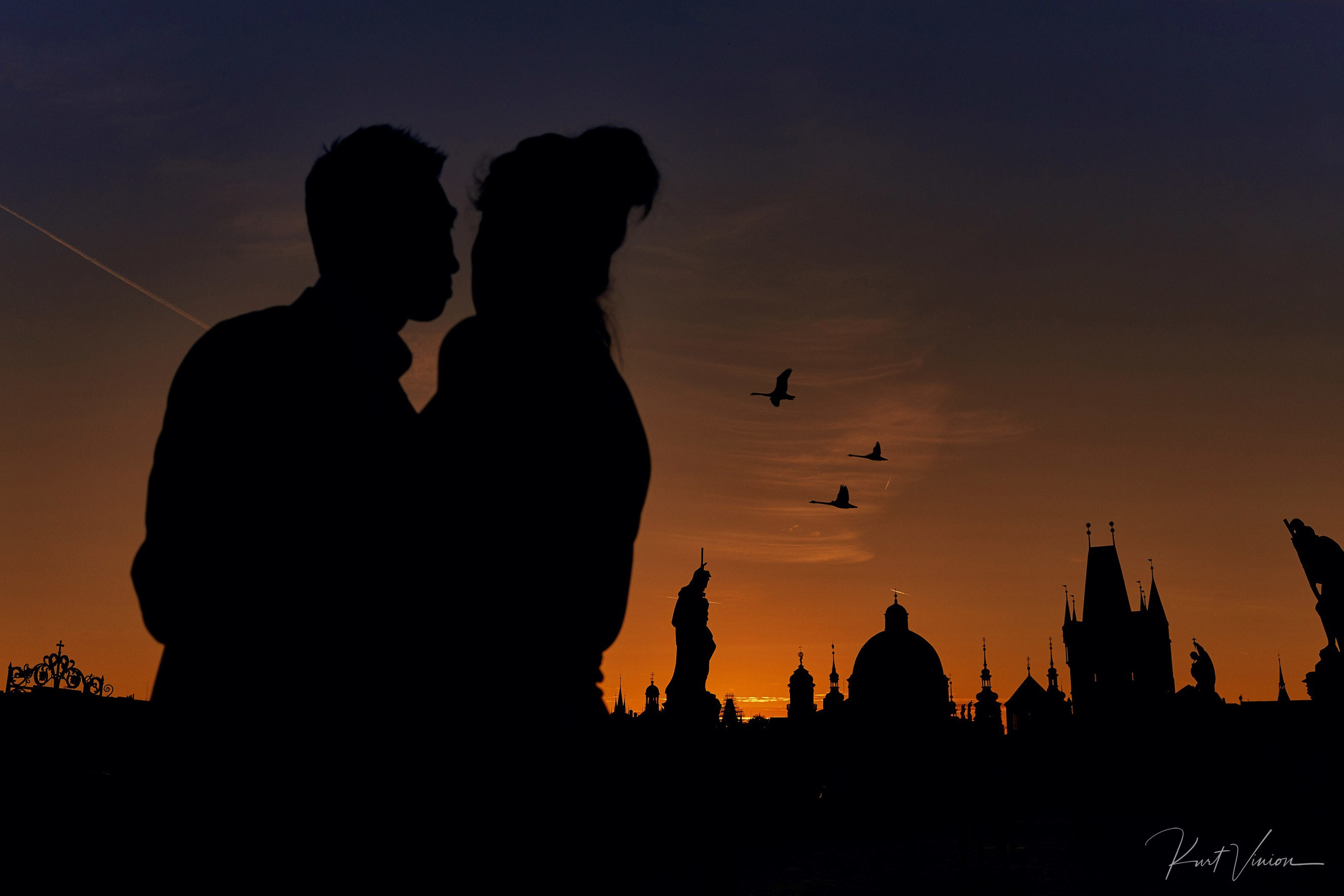 Silhouetted couple watching sunrise over Prague with swans moments after surprise marriage proposal on Charles Bridge