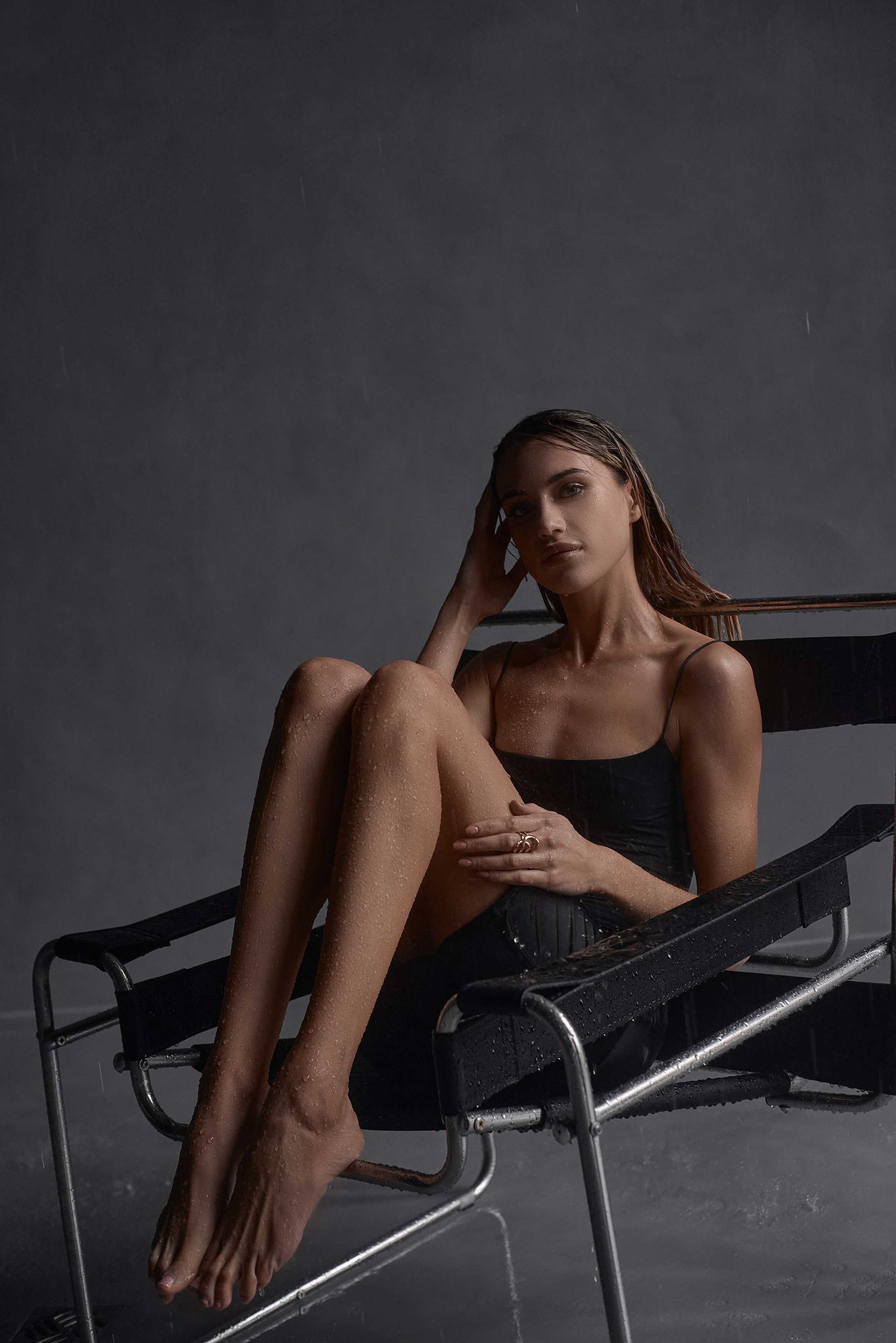 Молода жінка в чорній сукні сидить на кріслі у фотостудії під дощем, передаючи відчуття релаксації та ніжності.