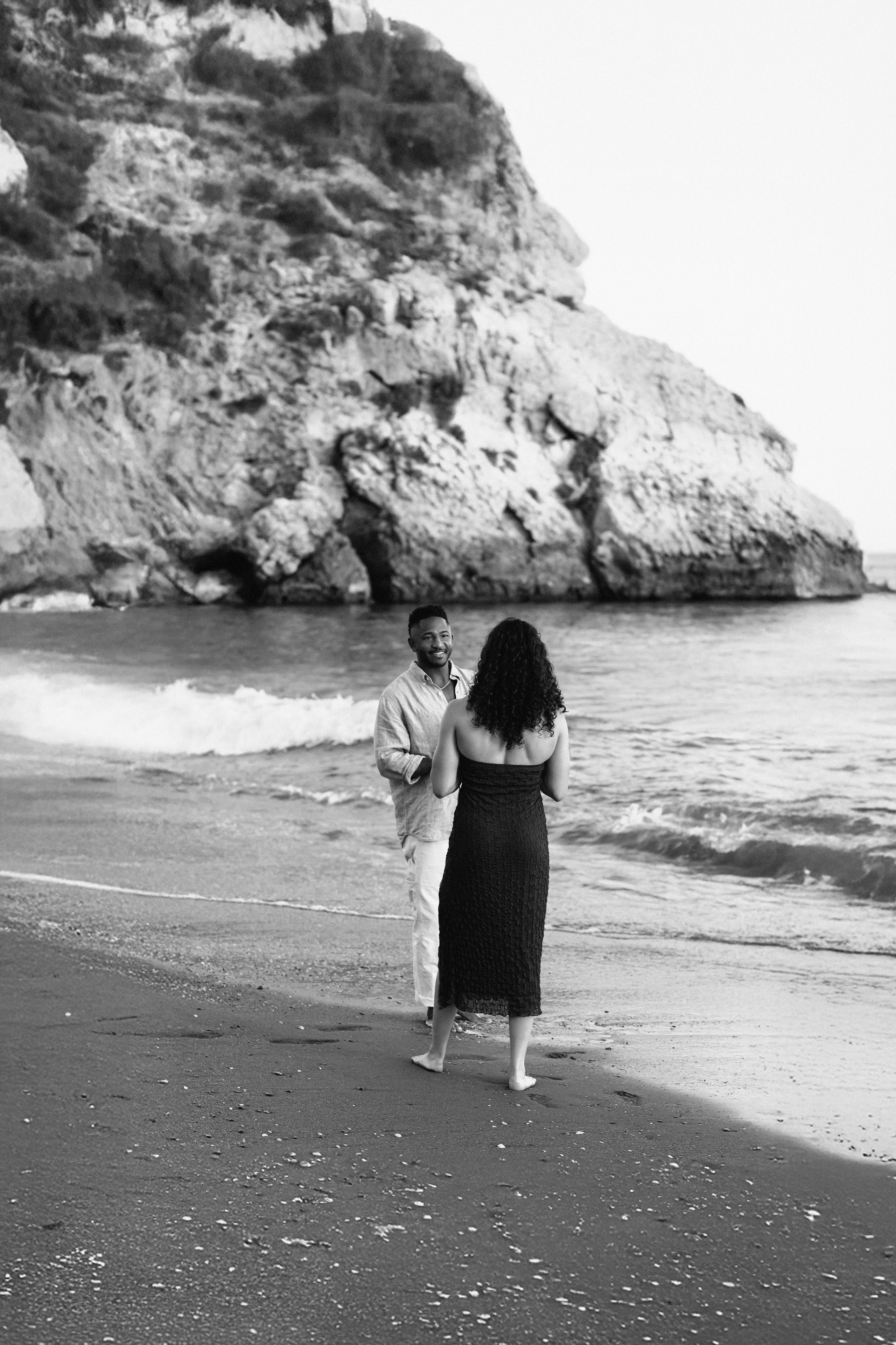 Black and white destination wedding engagement photo in Málaga, Spain, showing couple walking along the beach with dramatic coastal cliffs and the Mediterranean Sea in the background.