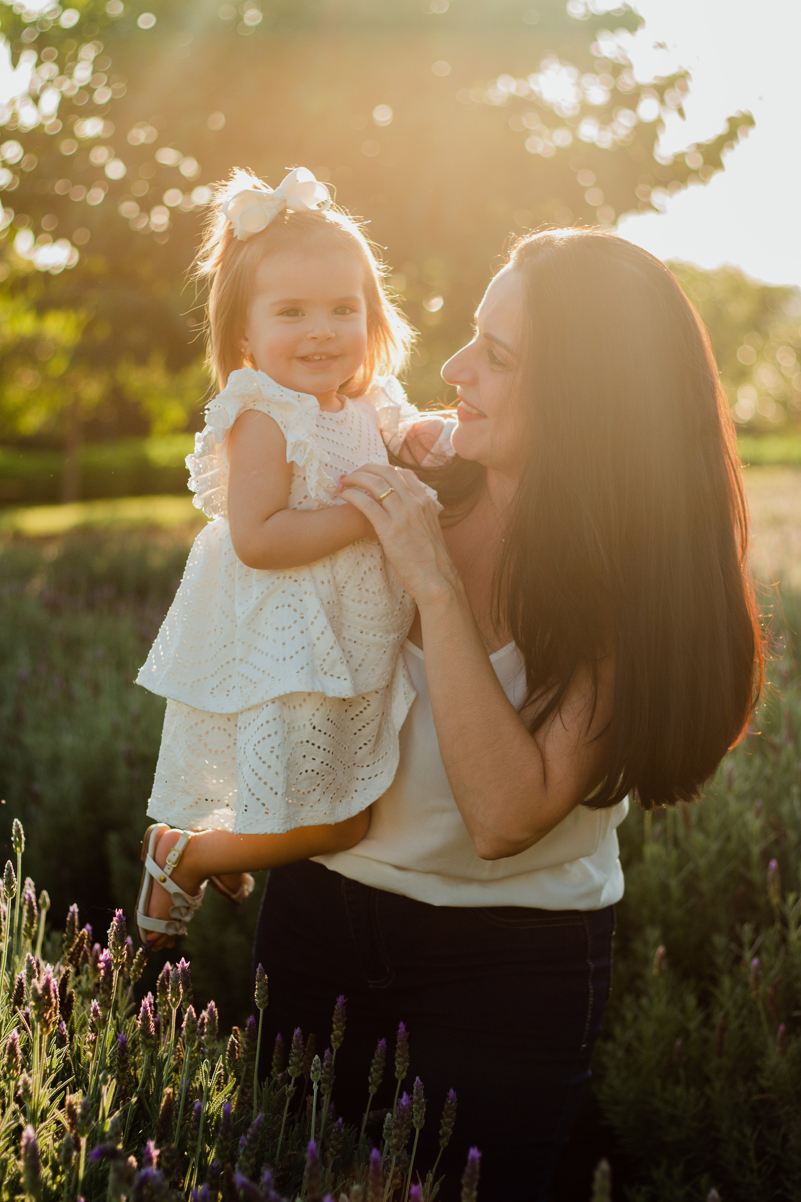 Ensaio de família em Holambra com mãe e filha usando roupas brancas, brincando entre lavandas ao entardecer, luz suave alaranjada do pôr do sol.