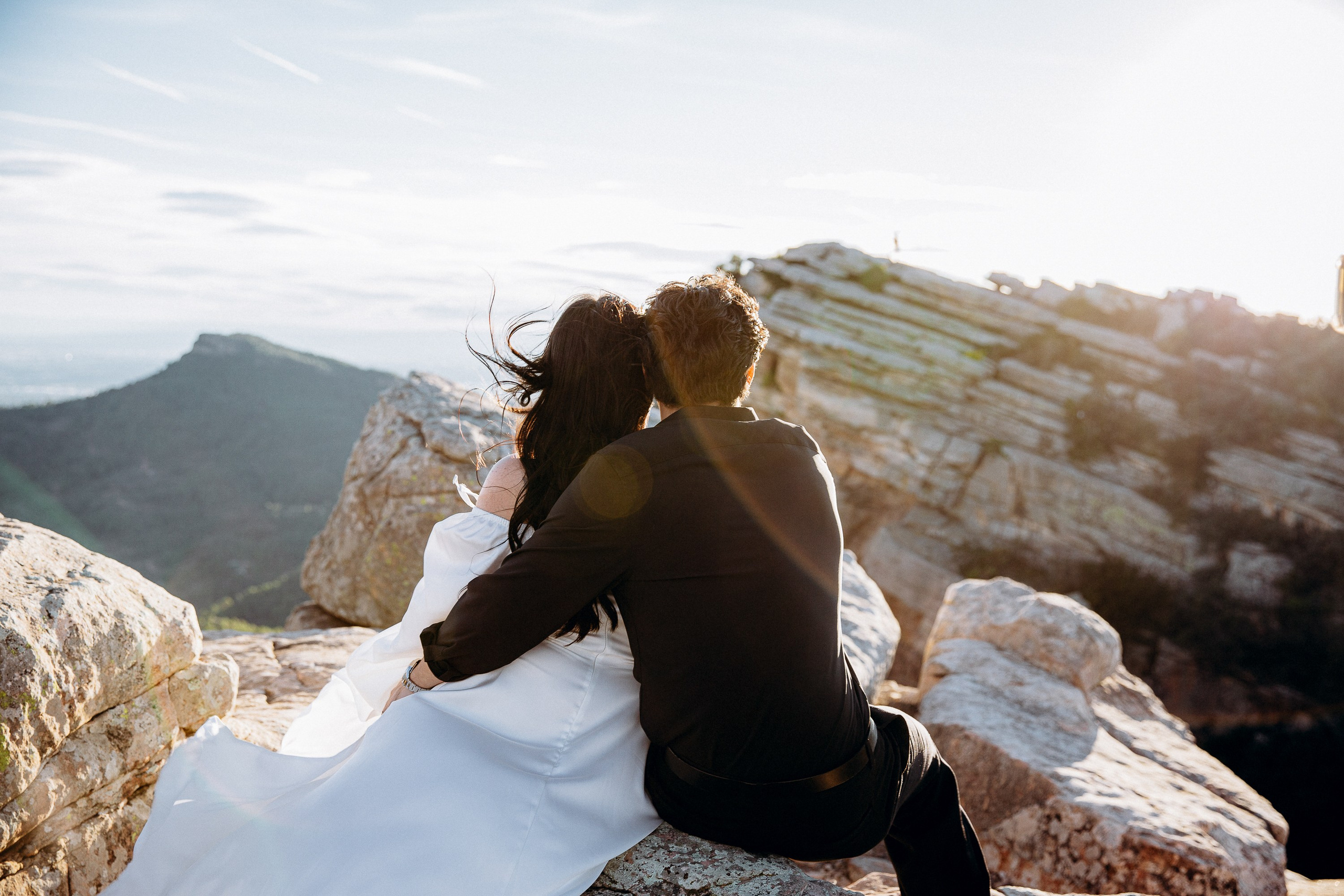 Engagement photoshoot in València, Spain, capturing a couple sitting closely together on rocky cliffs at sunset, overlooking a dramatic mountain landscape with soft golden light — a romantic engagement and wedding love story image ideal for engagement photography, pre-wedding sessions, elopements, and professional wedding photoshoots in València and across Spain.