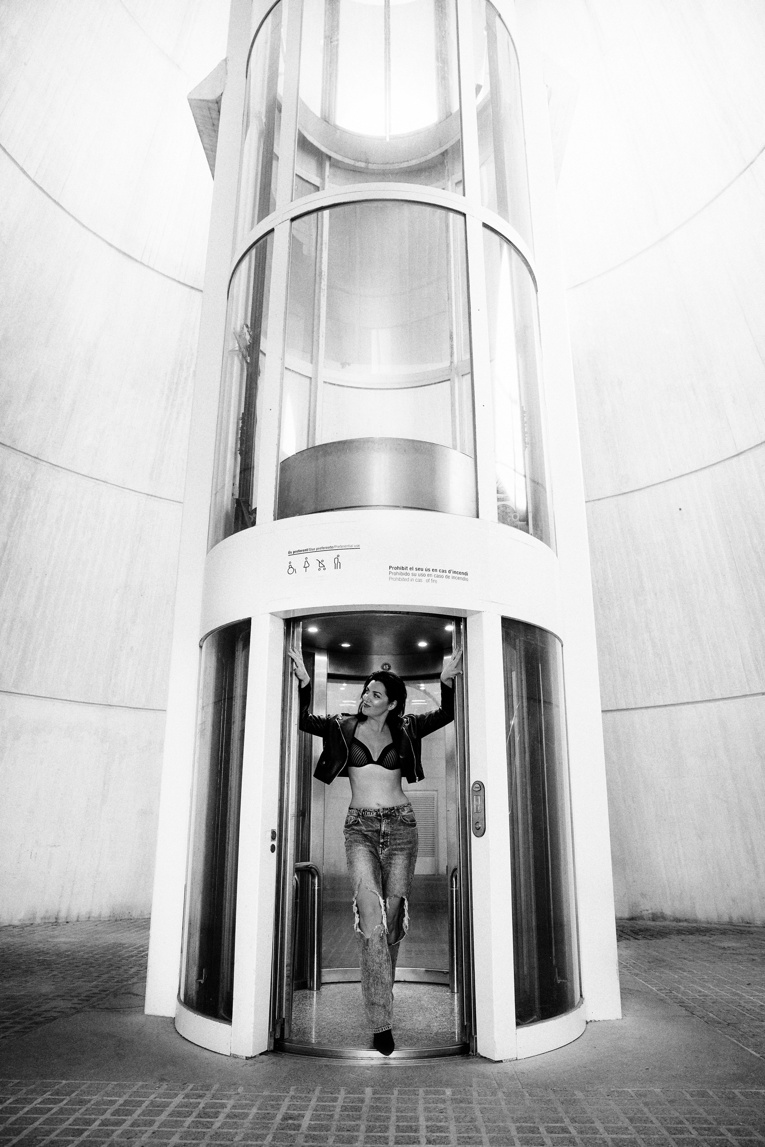 Черно-белый модный портрет уверенной женщины в укороченном топе, рваных джинсах и на каблуках, позирующей в дверном проеме современного стеклянного лифта в Валенсии, Испания