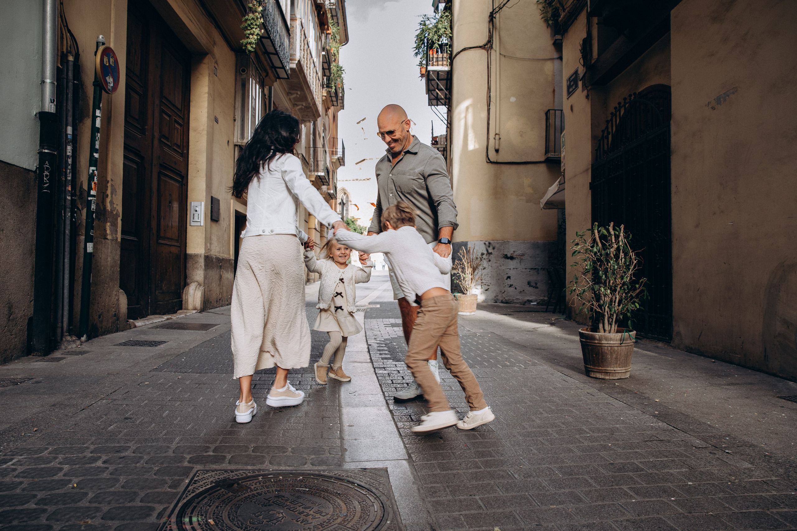 Sesión de fotos familiar en Valencia, España, con unos padres felices jugando con su pequeña hija mientras la sostienen de las manos en una encantadora calle del casco histórico rodeada de arquitectura mediterránea. Un retrato familiar lifestyle espontáneo que transmite alegría, movimiento y una auténtica conexión familiar. Ideal para fotografía familiar, sesiones de fotos familiares, retratos infantiles, fotografía de vacaciones y fotografía familiar de destino en Valencia y toda España.