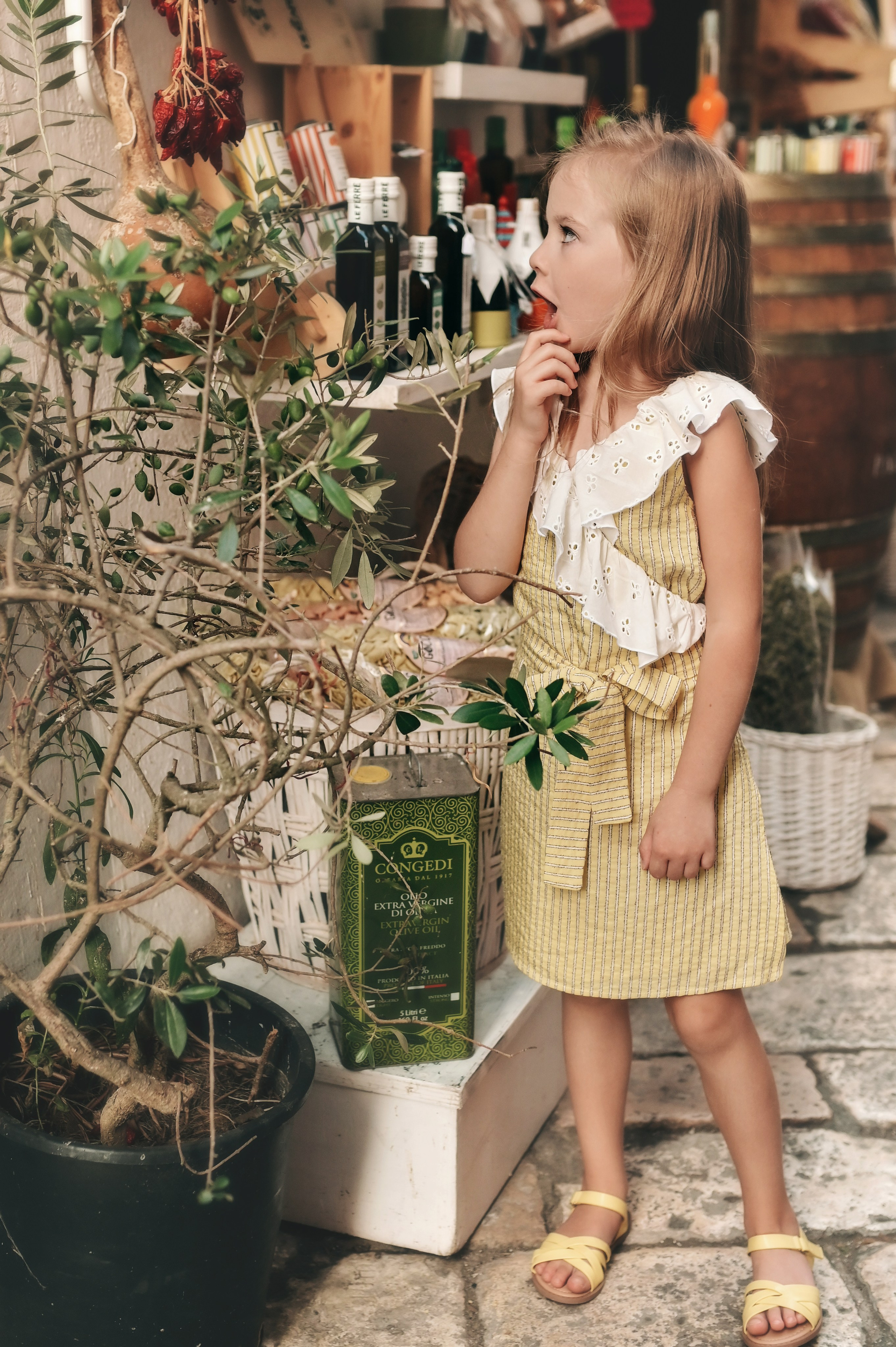 A young girl smiling while reaching for dried chili peppers in a local shop filled with bottles of olive oil and other Italian products.