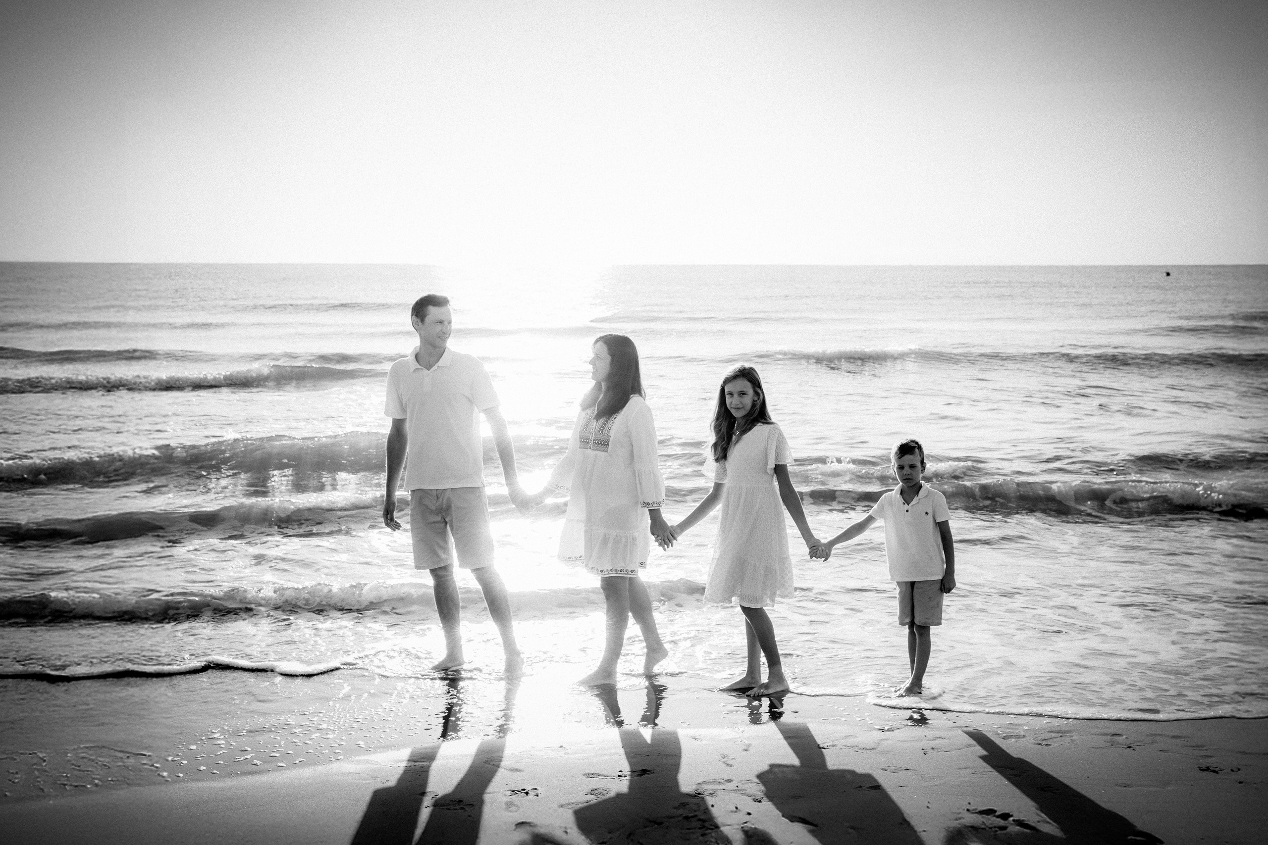 Sesión familiar en la playa en blanco y negro en Valencia, España, con padres e hijos tomados de la mano junto a la orilla — perfecta para quienes buscan fotografía familiar elegante y atemporal en Valencia y España. Ideal para retratos artísticos junto al Mediterráneo.