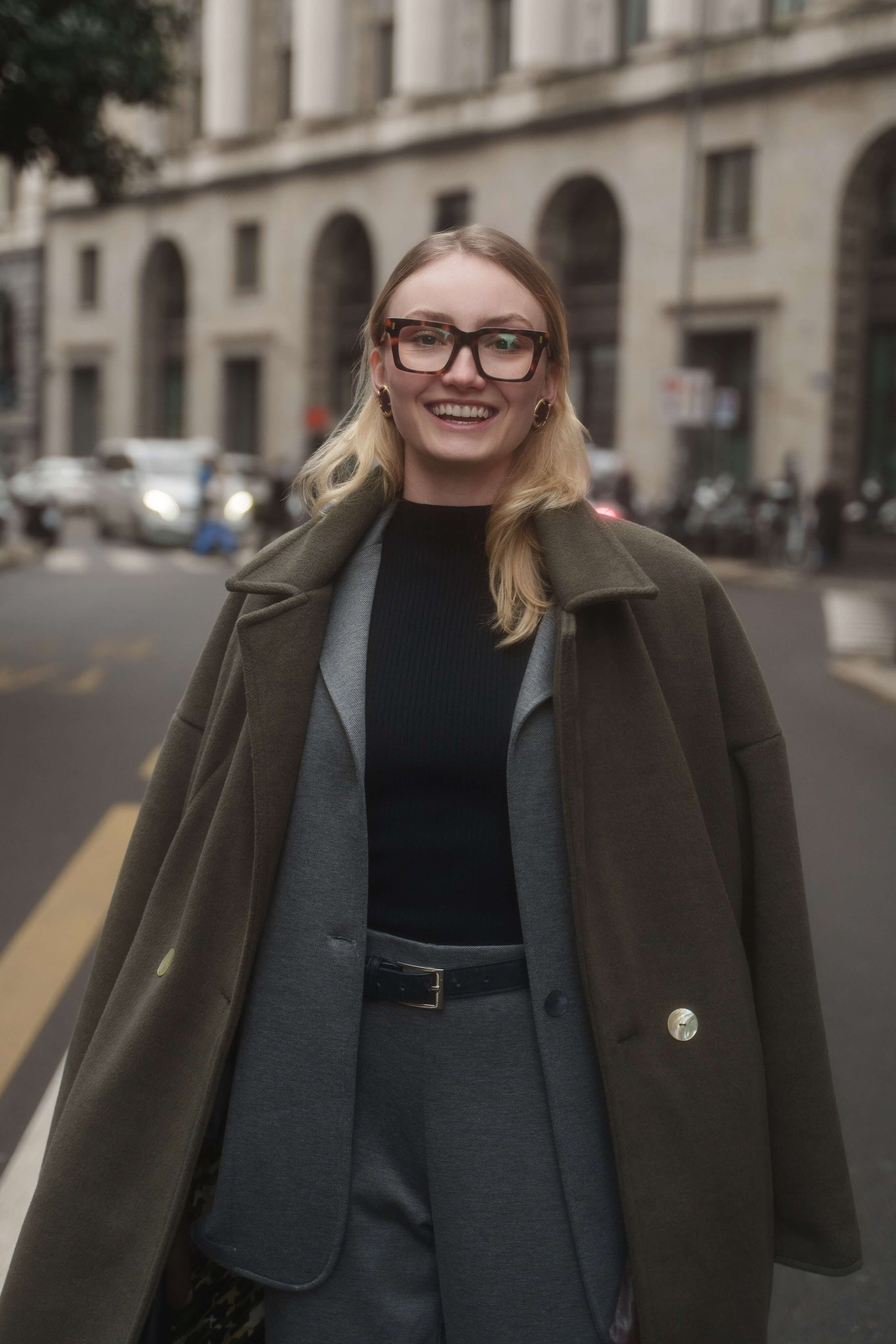 Smiling woman in oversized tortoiseshell glasses wearing an olive green coat on a Milan street