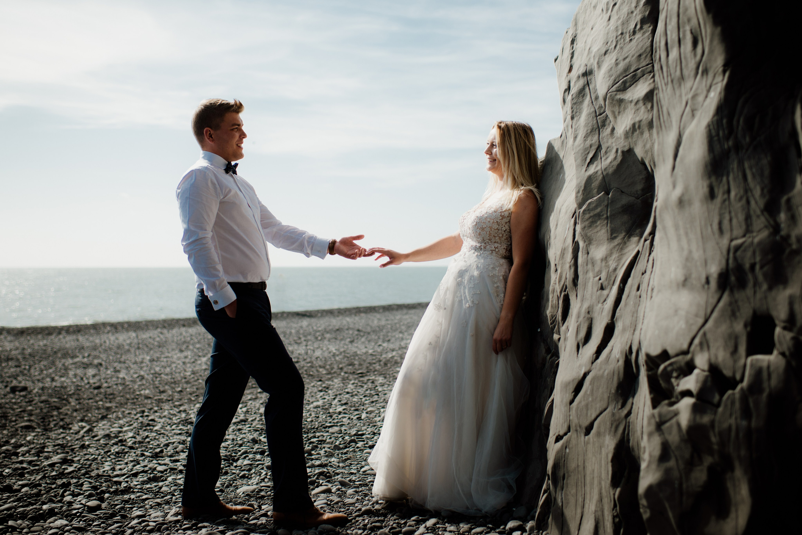 Bride in a flowing dress standing on a cliffside, with the groom gazing at her lovingly.