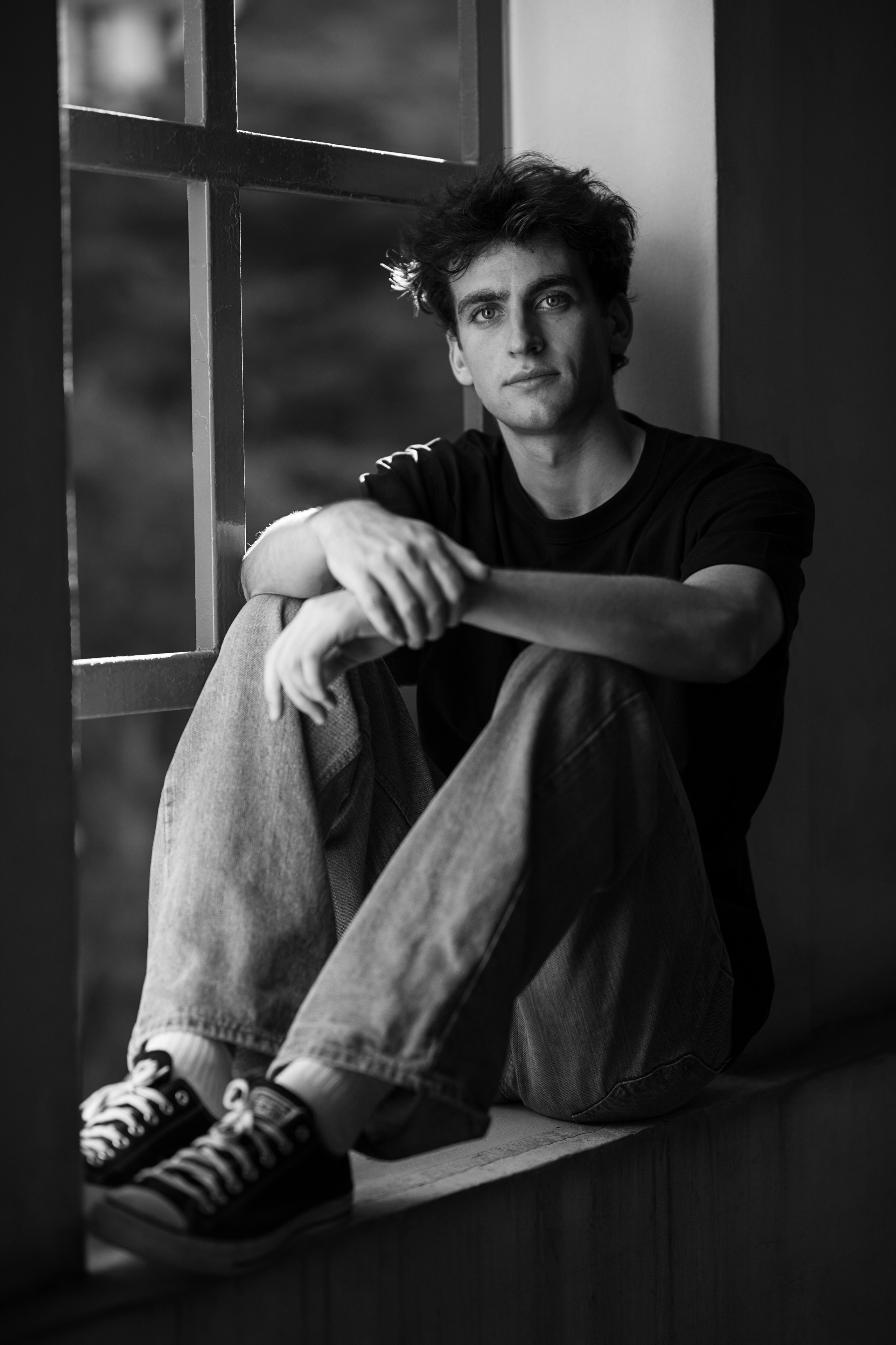 Monochrome portrait of a man seated on a window ledge in soft window light, jeans and sneakers visible; photographed in Singapore.