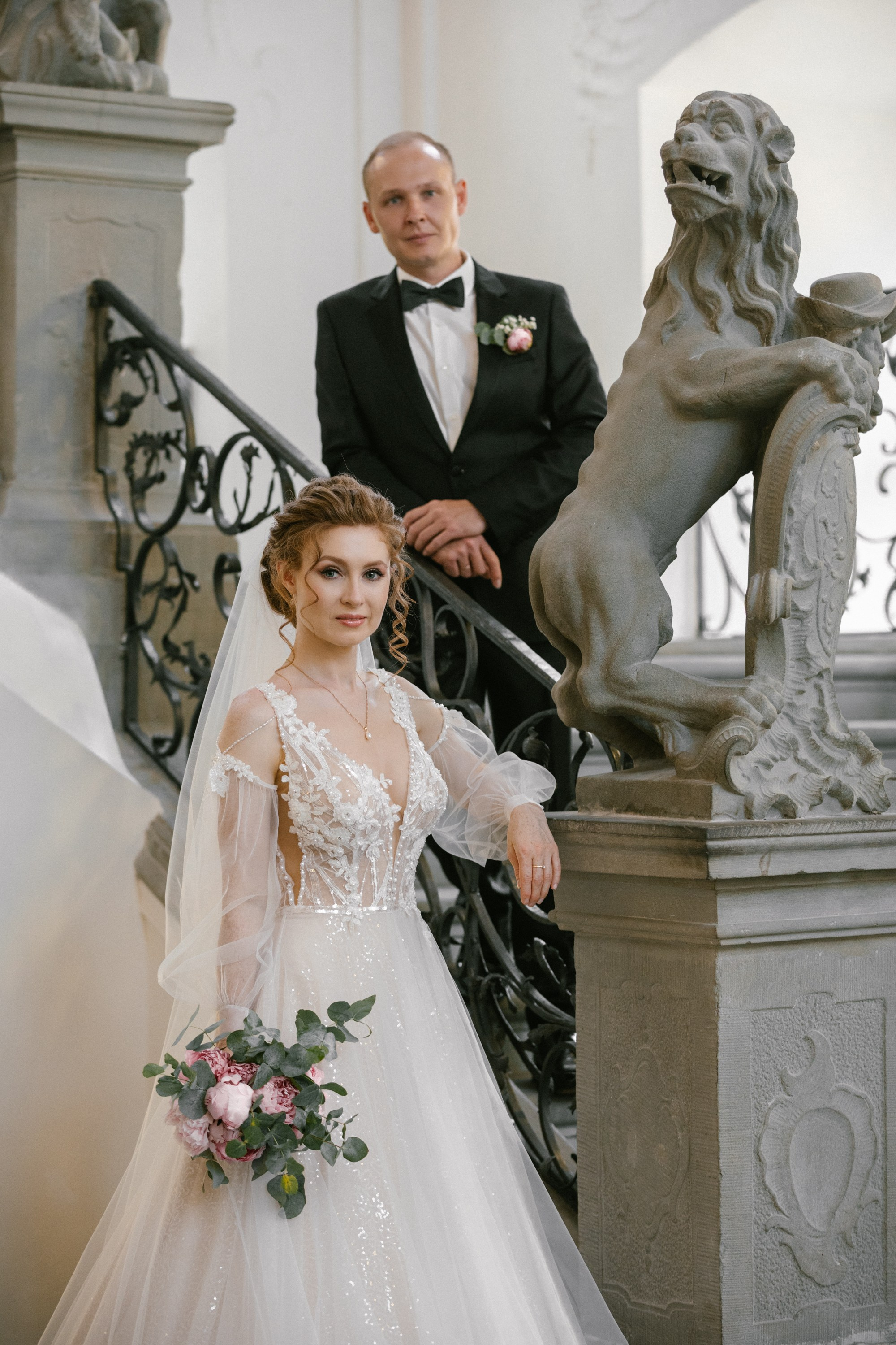 Bride stands by stone lion sculpture holding bouquet, groom above on stairs at Schloss Meersburg