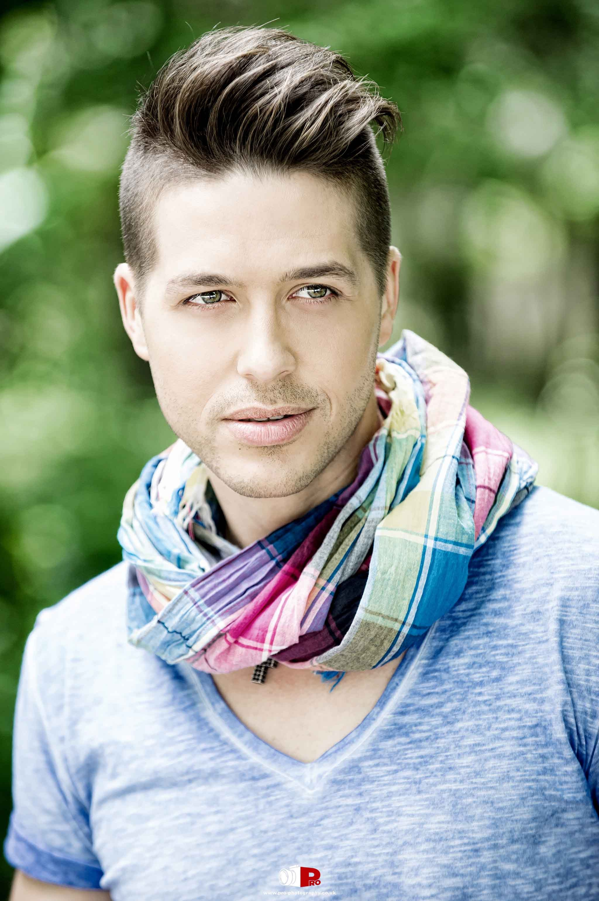 Relaxed portrait of a stylish young man with a trendy hairstyle, wearing a light blue t-shirt and colourful scarf, posing outdoors in natural surroundings.