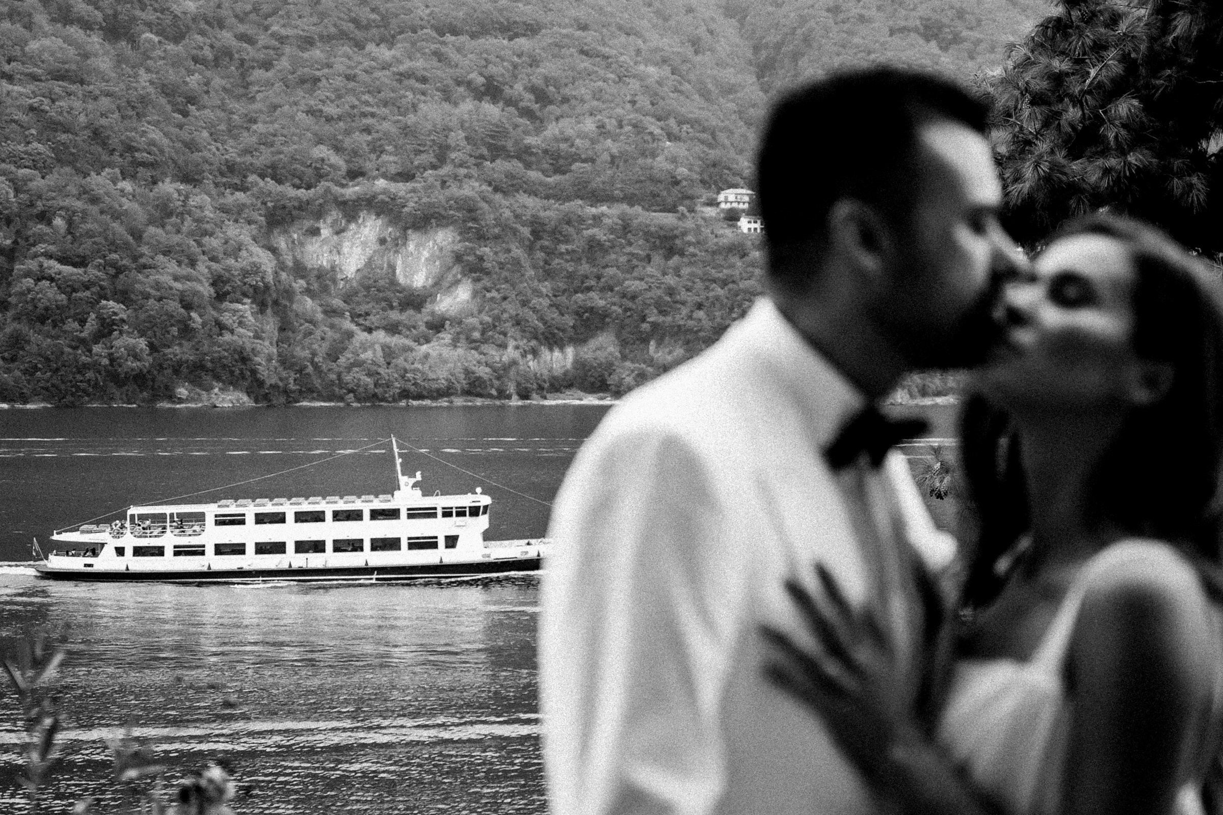 Black and white romantic photo of couple embracing by lakeside with ferry and forested hills in the background.