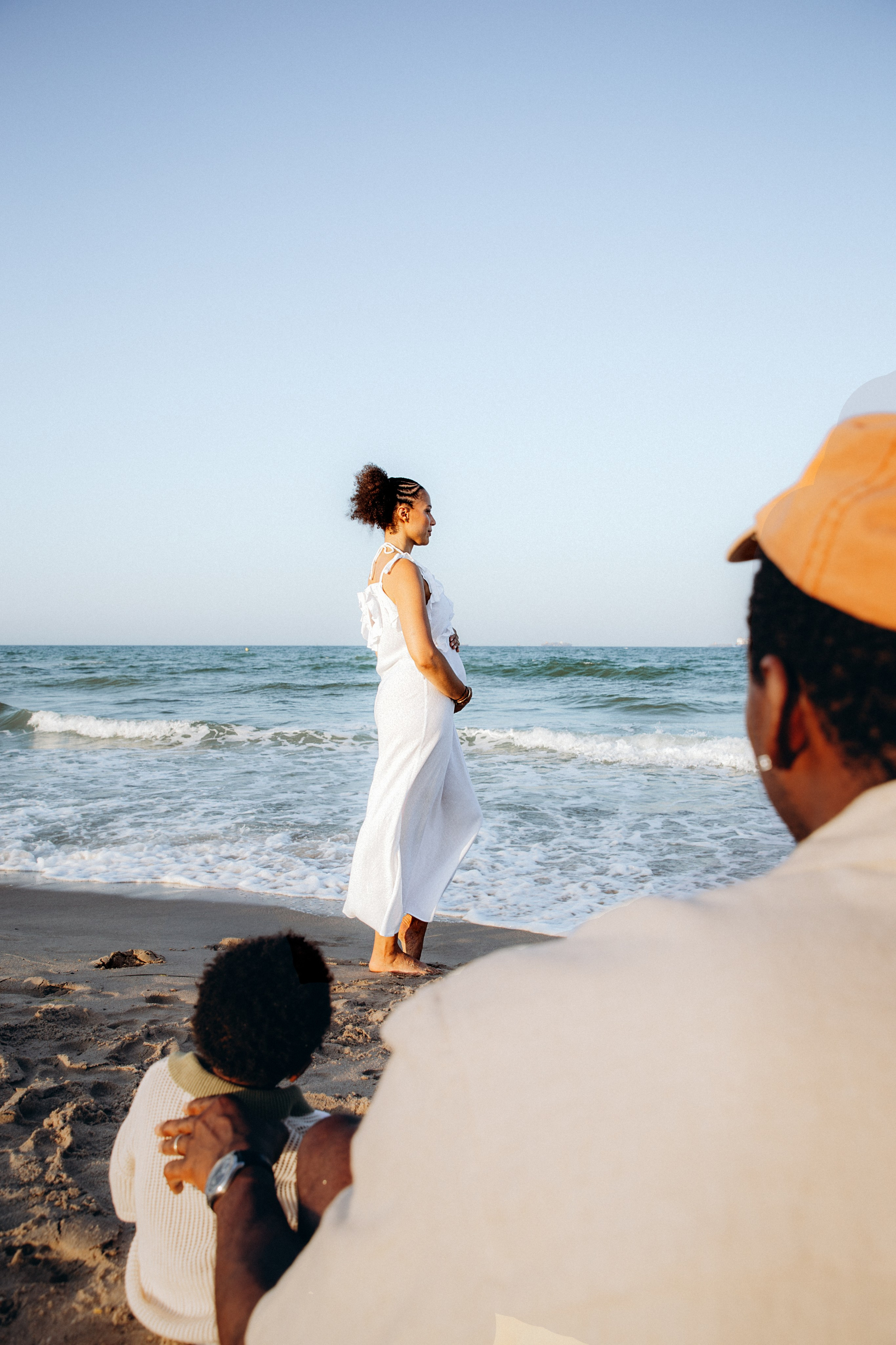 Sesión espontánea de embarazo en la playa de Barcelona, España — mujer embarazada con vestido blanco posando descalza junto al mar, mientras su pareja e hijo la observan desde la distancia. Inspiración para fotografía natural de embarazo en la costa catalana.