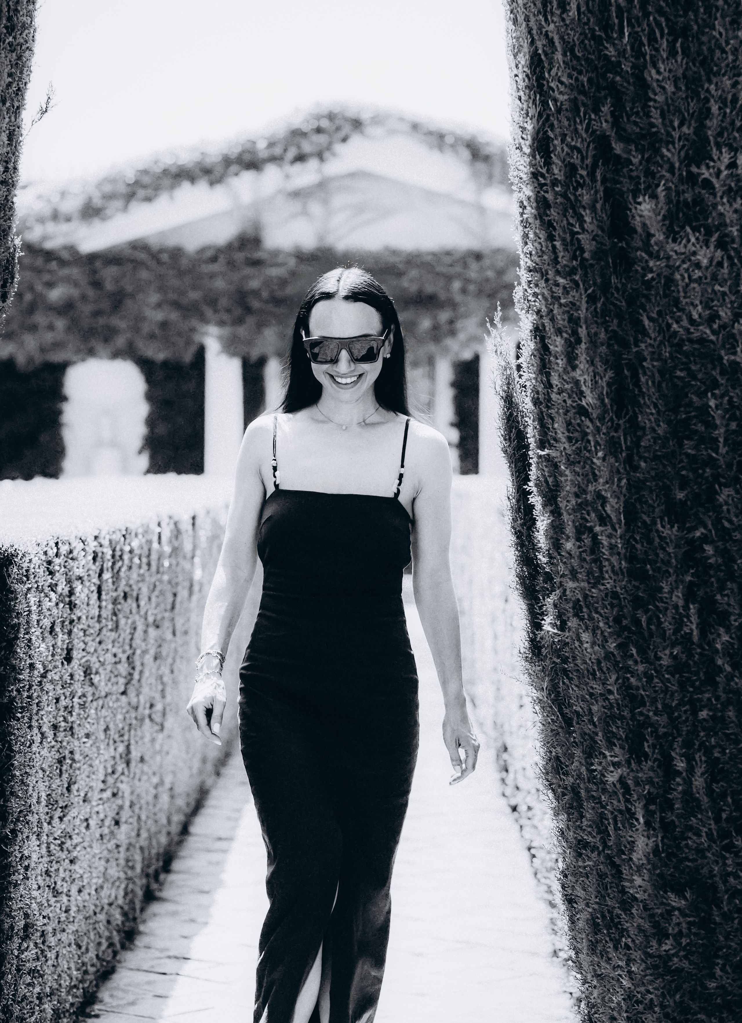 Стильный черно-белый портрет женщины в строгом черном платье и солнечных очках, идущей по садовой аллее в Дении, Испания — отличное вдохновение для модных и современных портретных фотосессий в Дении и на побережье Коста-Бланка.