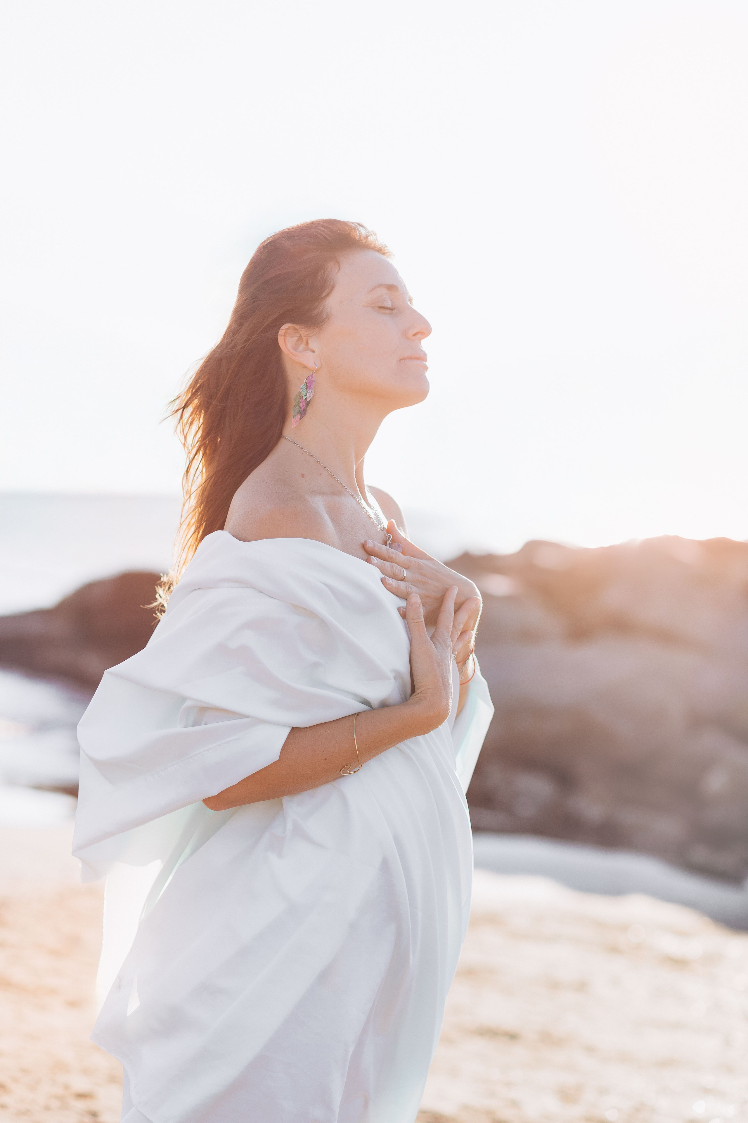 Спокойный портрет беременной женщины на пляже в воздушной белой накидке, с закрытыми глазами и руками на груди, освещённой мягким золотым светом, на фоне скалистого берега в Валенсии, Испания