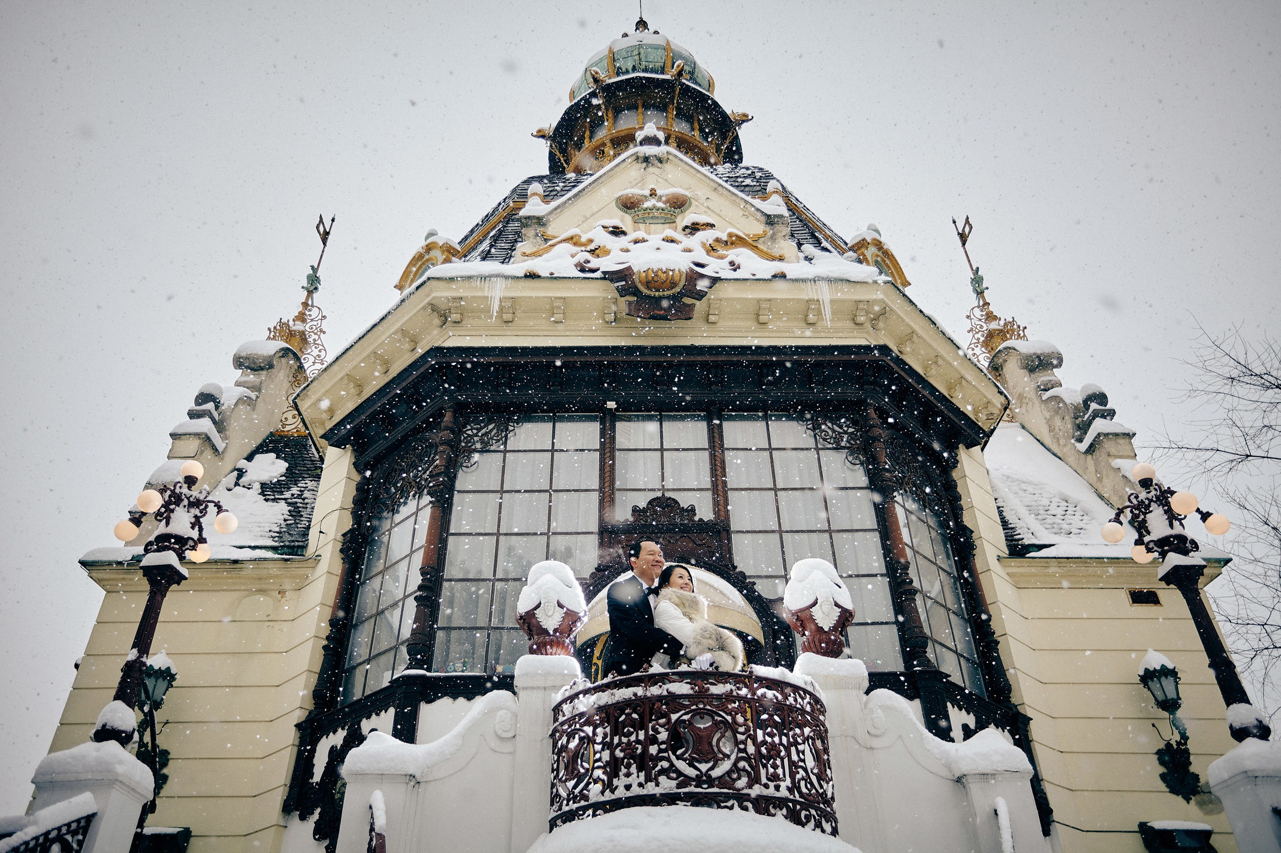 Older, distinguished looking Asian man cuddles with his beautiful partner as they view the winter sceneryof Prague from the balcony of the Hanavský pavilon as snow falls
