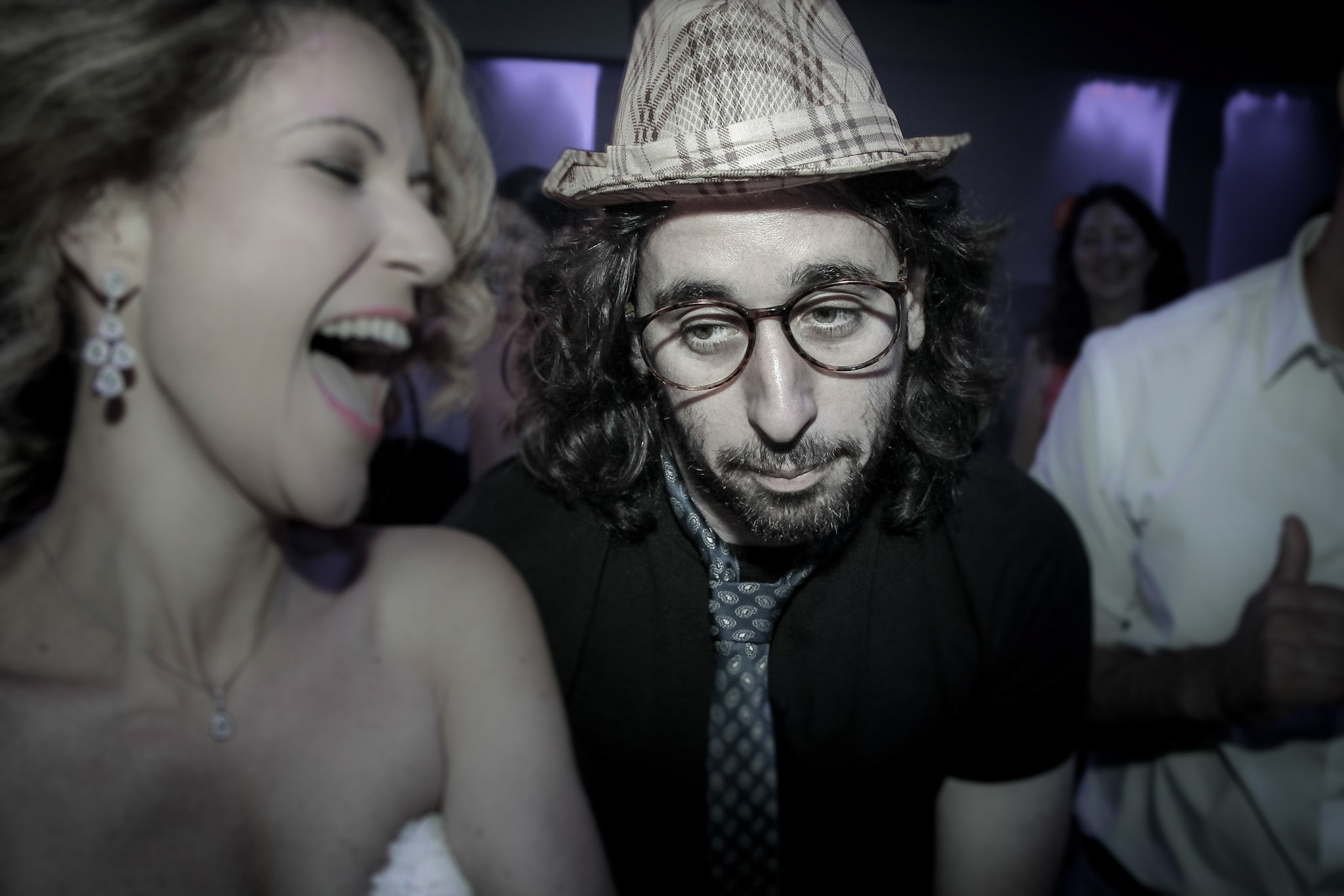 A photo capturing contrasting emotions at a wedding party: a bride in a white dress laughs joyfully in the foreground, while a man in a hat and glasses looks serious and thoughtful in the blurred background, hinting at the festive atmosphere. The image contrasts carefree joy with introspective seriousness, emphasizing the different emotional states present in the same moment at the event.