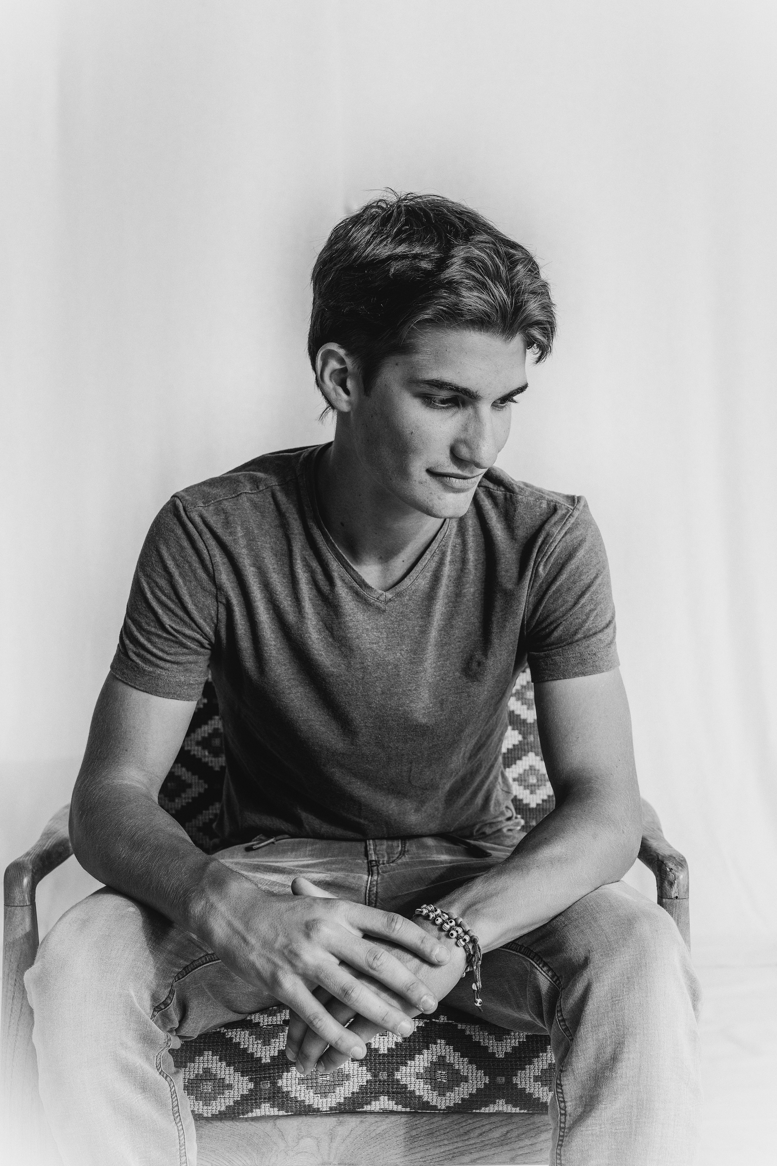 Black and white portrait of a young man sitting in a chair, wearing a casual t-shirt and jeans, with a thoughtful expression. Ideal for those seeking professional portrait photography that captures natural and introspective moments. Portrait en noir et blanc d'un jeune homme assis sur une chaise, portant un t-shirt décontracté et un jean, avec une expression pensive. Idéal pour ceux qui recherchent des services de photographie de portrait professionnelle capturant des moments naturels et introspectifs.