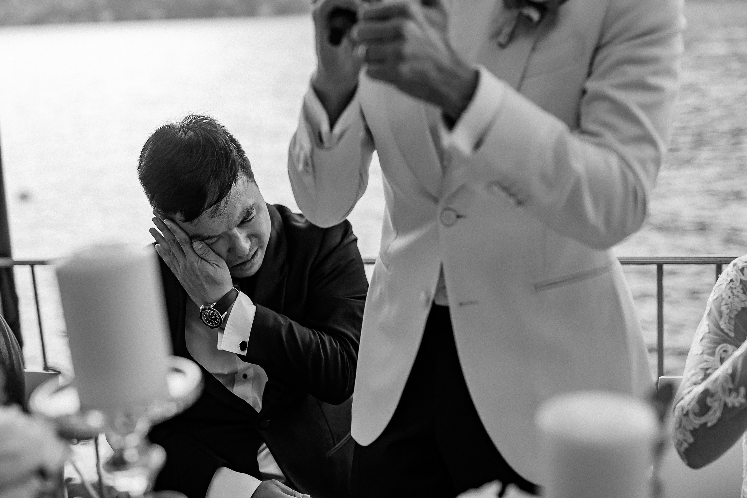Man in a suit wipes tears while another stands in a white jacket at a lakeside dinner.