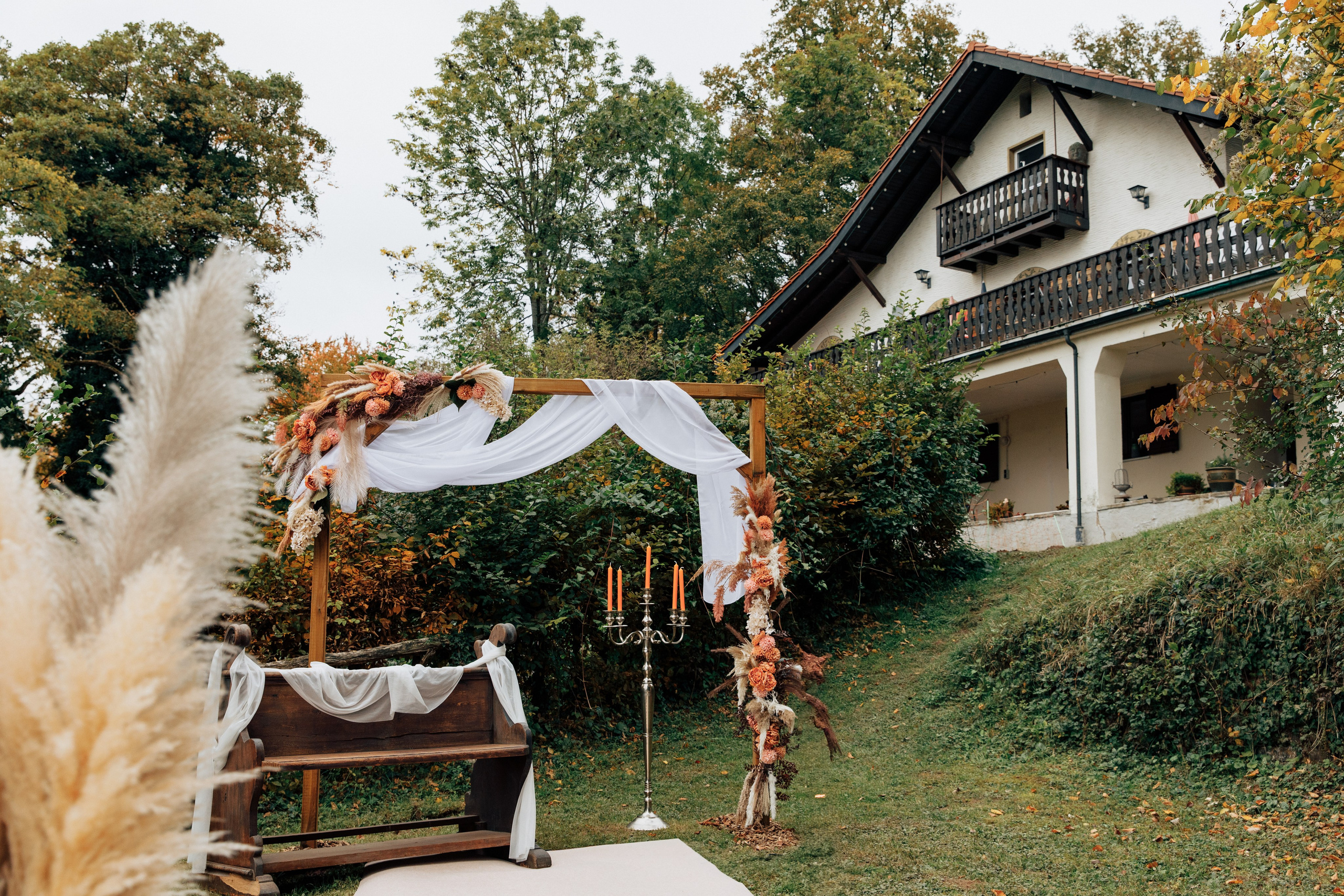 Im Vordergrund steht eine antike Bank und ein Hochzeitsbogen, dekoriert mit Blumen und Pampasgras. Im Hintergrund ist das Gebäude des Restaurants Almhütte Kleinwallstadt zu sehen.
