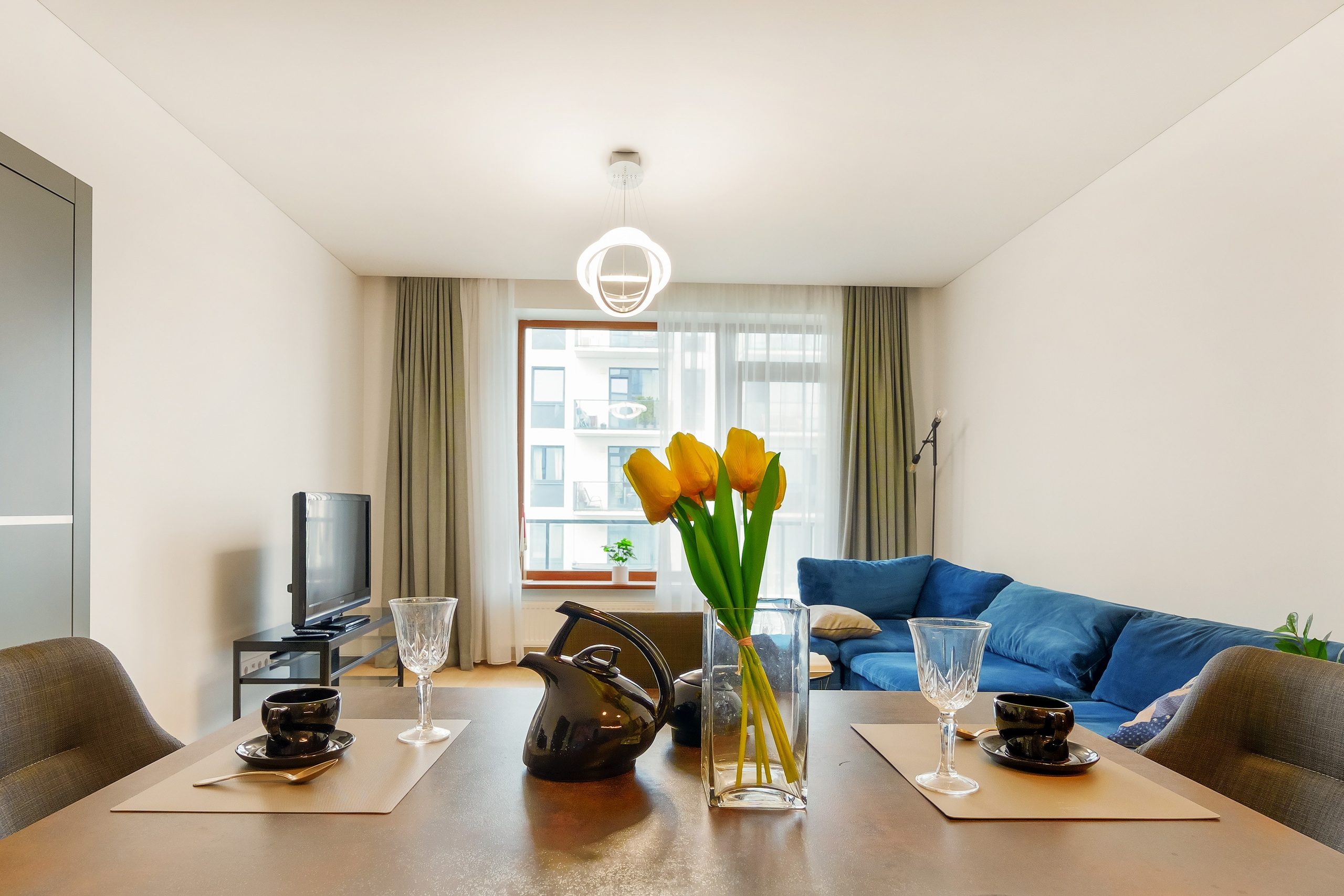 Moderni svetainė su valgomojo stalu dviem asmenims, geltonomis tulpėmis vazoje, mėlyna sofa, televizoriumi ir dideliu langu su užuolaidomis. – nuostabiai užfiksavo nt fotografas sapiegafoto.lt