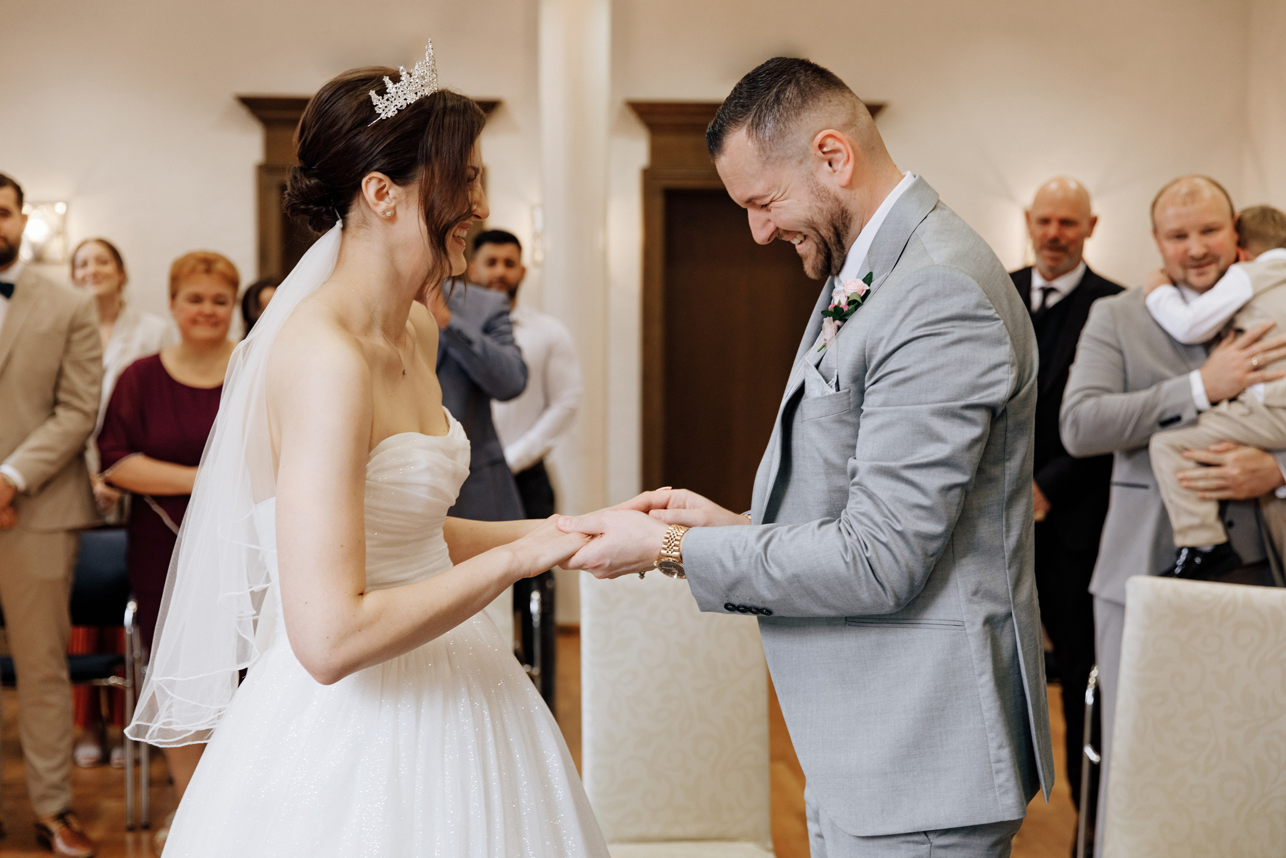 Der Bräutigam zieht der Braut den Ehering an, während sie lachen und die Gäste ebenfalls lächeln. 