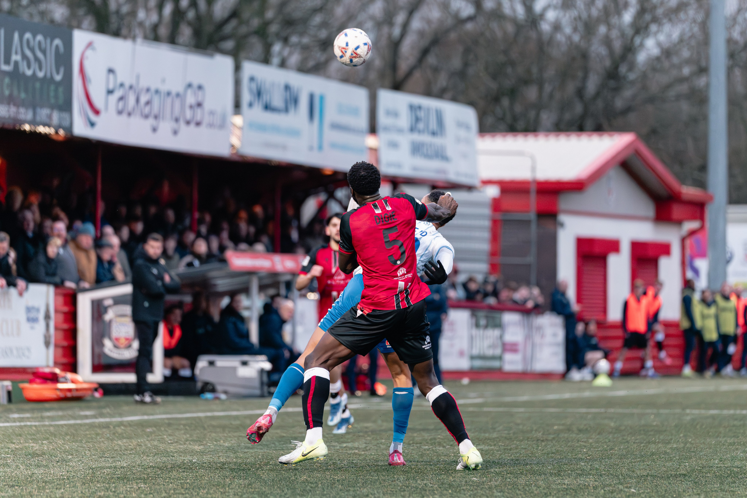 Kennedy Digie in a red Tamworth FC shirt jumps into an aerial duel with a Hartlepool United F.C. player in blue and white as the ball drops above them, with the stand and spectators in the background.