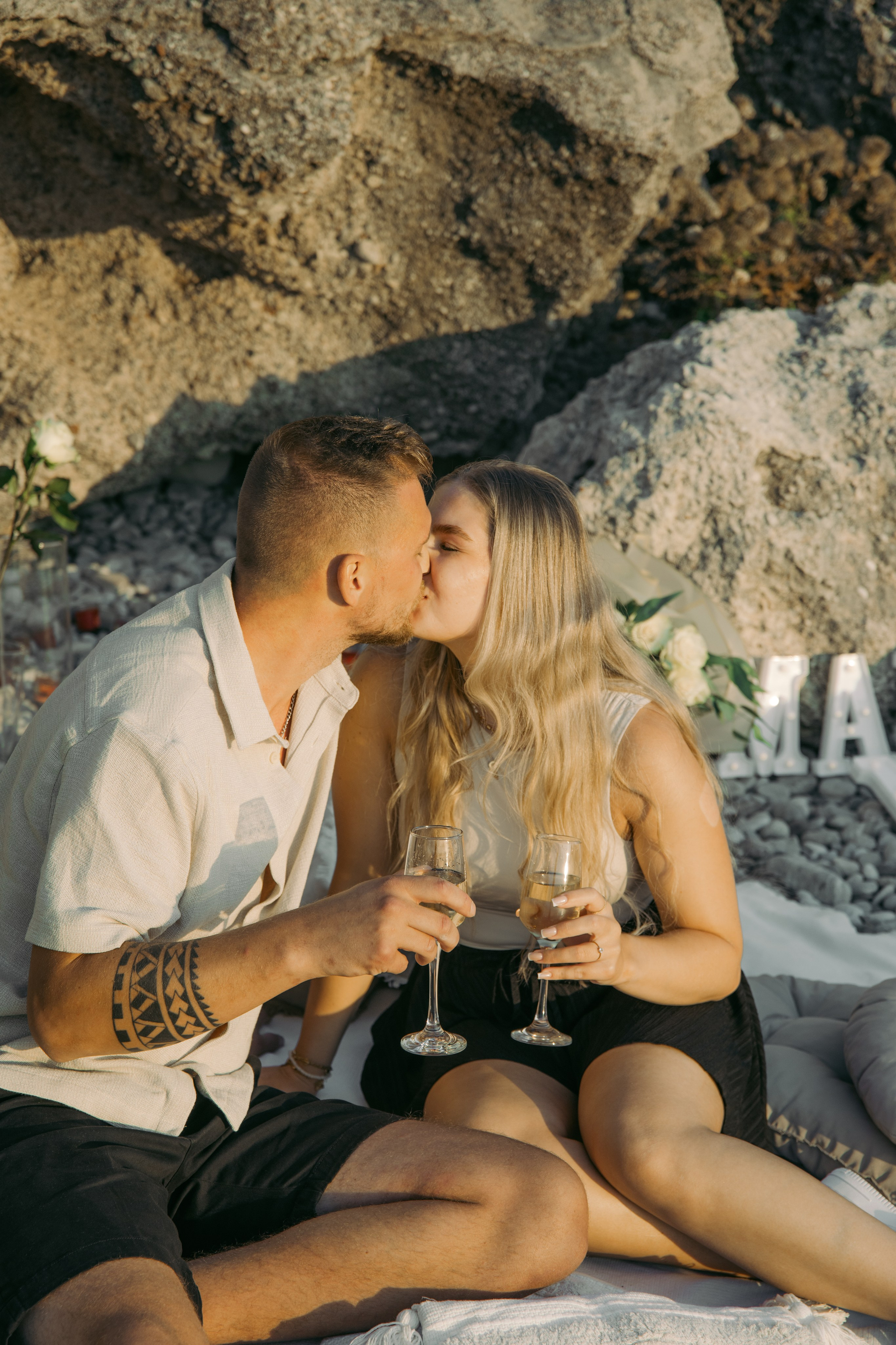 "Nahaufnahme eines Paares bei einem zärtlichen Kuss nach der Verlobung. Die Augen sind geschlossen, die Gesichter einander zugewandt, während die raue Brise der Ägäis und das Rauschen der Wellen im Hintergrund die friedliche Intimität dieses Augenblicks unterstreichen."