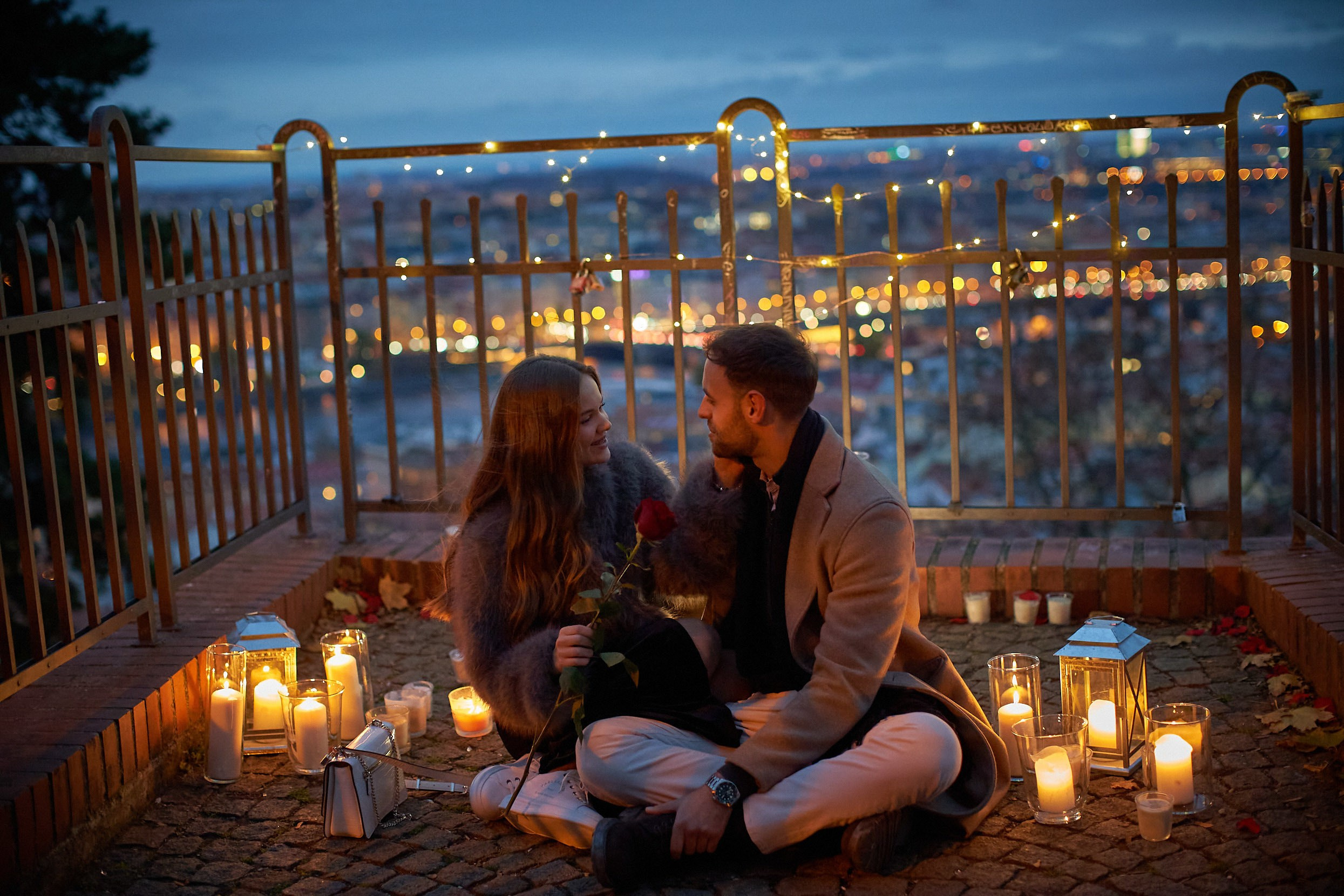 Woman holding rose sitting close to partner surrounded by candles and rose petals above Prague city lights.
