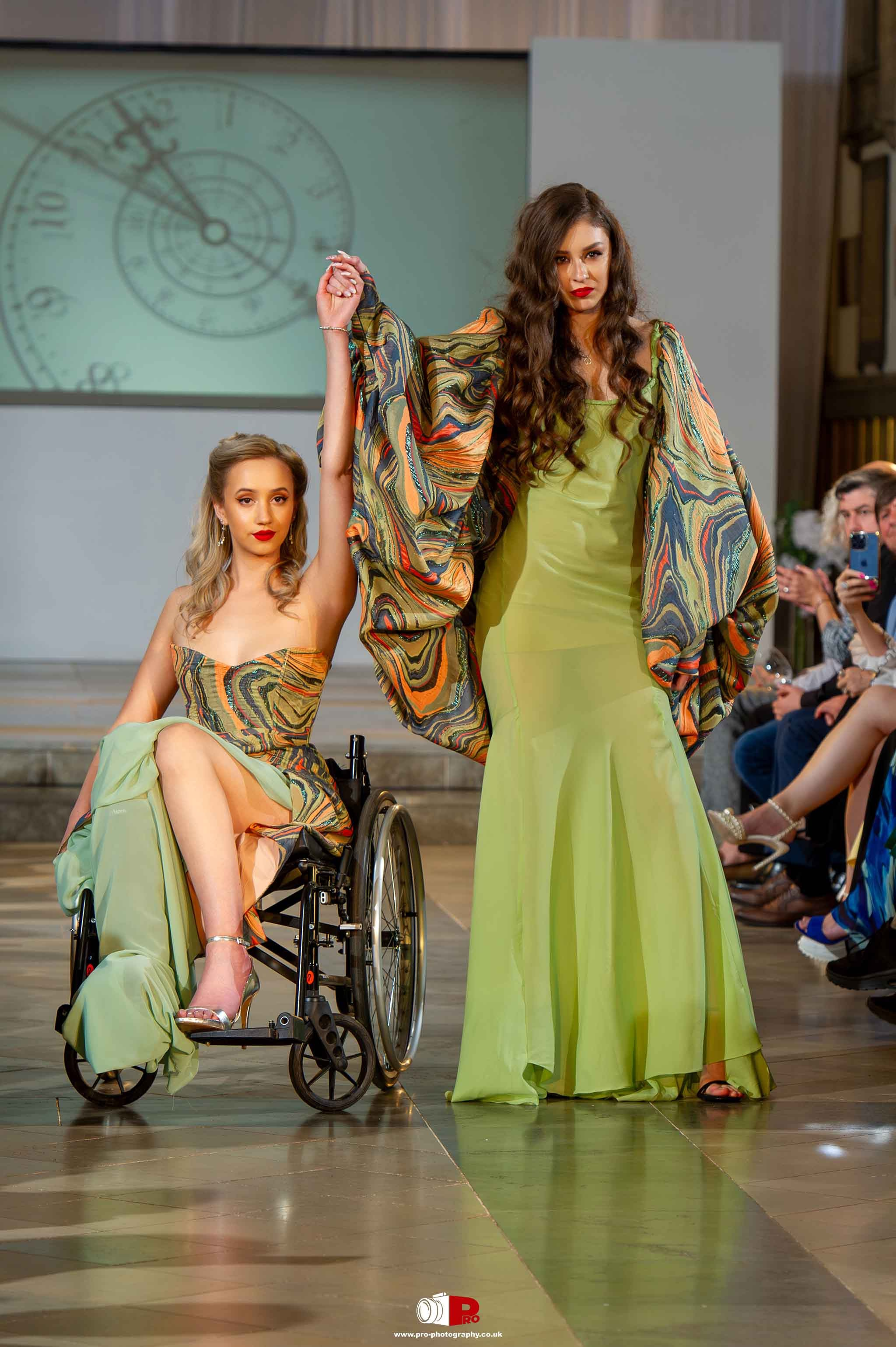 Two models on the runway, one in a wheelchair and one standing, wearing coordinated green and marbled outfits during a fashion show.