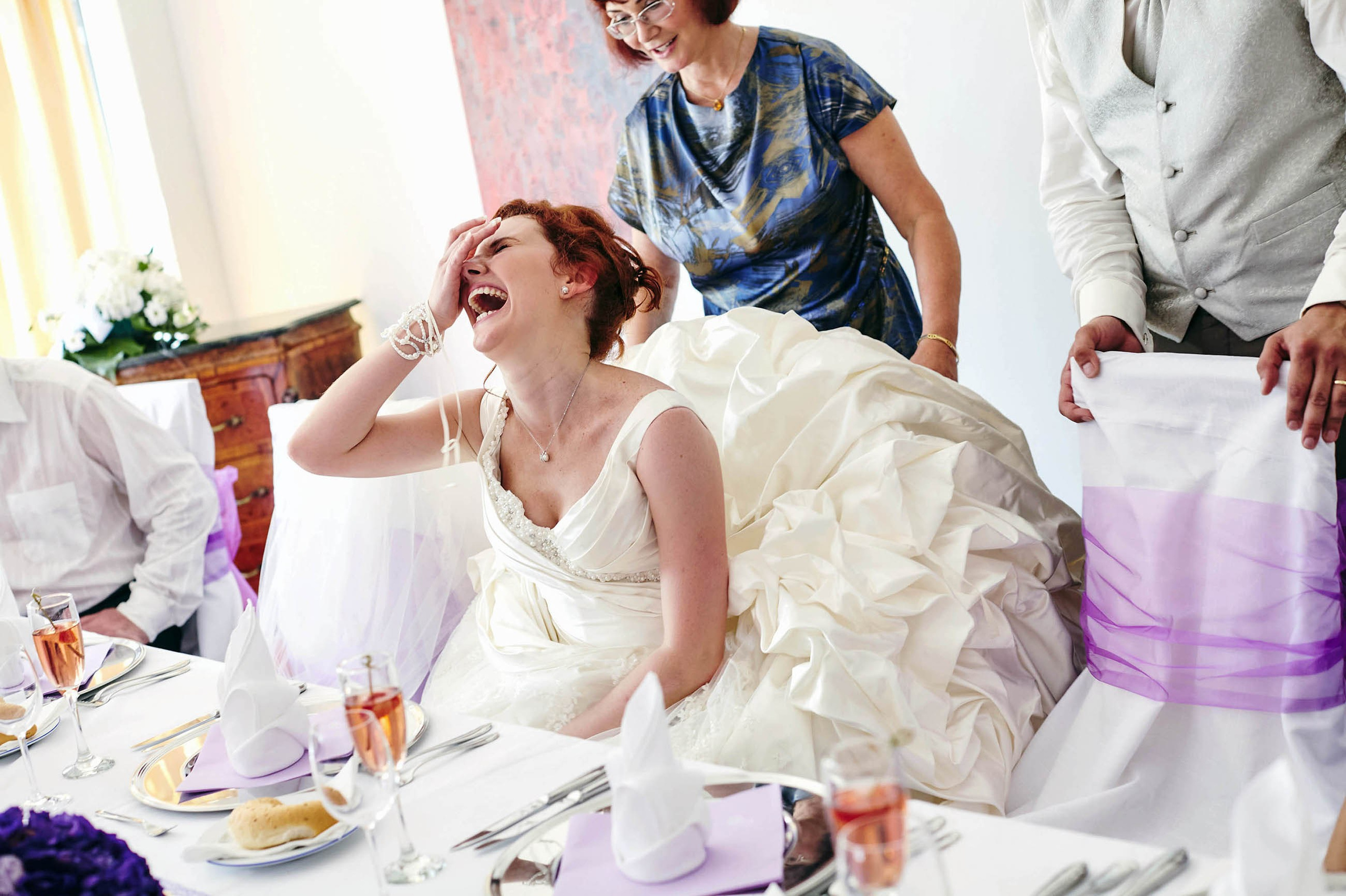 A laughing bride touches her face as she sits down in her large wedding dress as her mother assists during her wedding dinner.