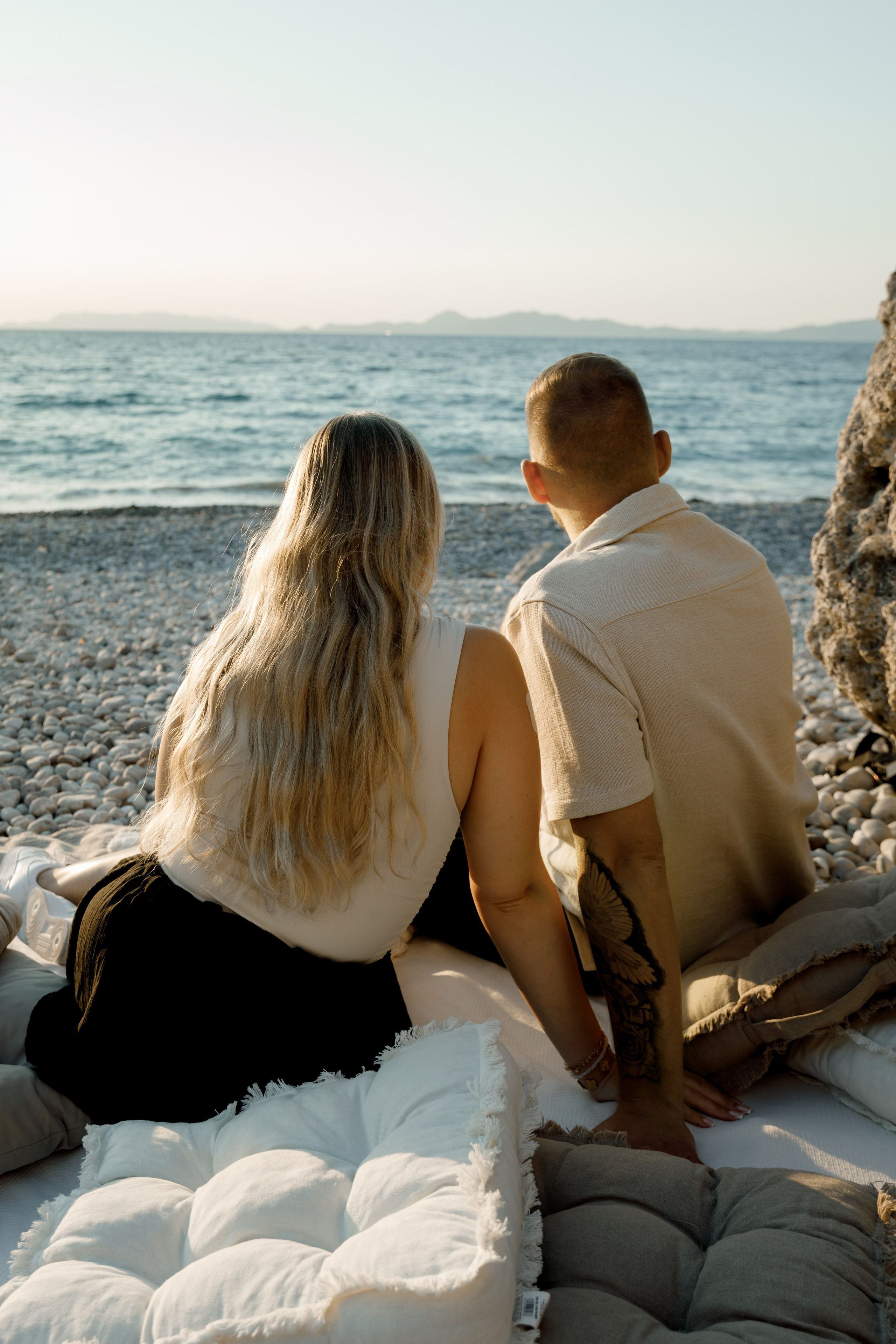 "Moment der Ruhe nach dem Ja-Wort: Ein Paar sitzt auf dem Kieselstrand von Kato Petres und genießt die Zweisamkeit. Die Wellen schlagen sanft an das steinige Ufer, und das warme Licht des Sonnenuntergangs taucht die Szenerie in goldene Farben."