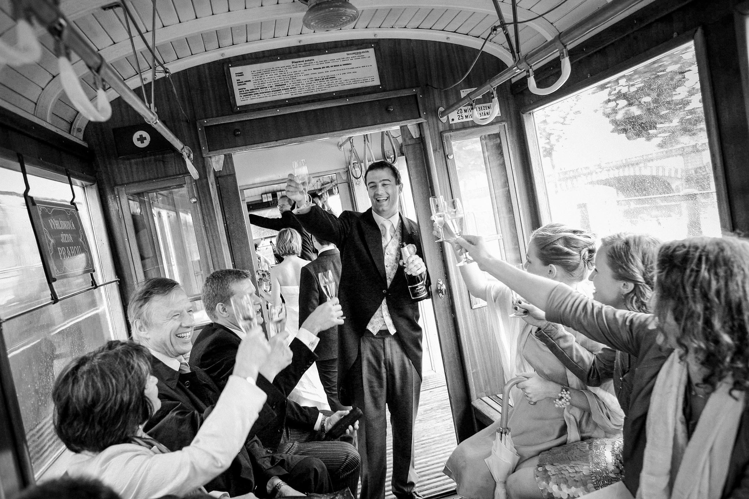 Black and white photo of English groom receiving toast on historical Prague tram with wedding party.